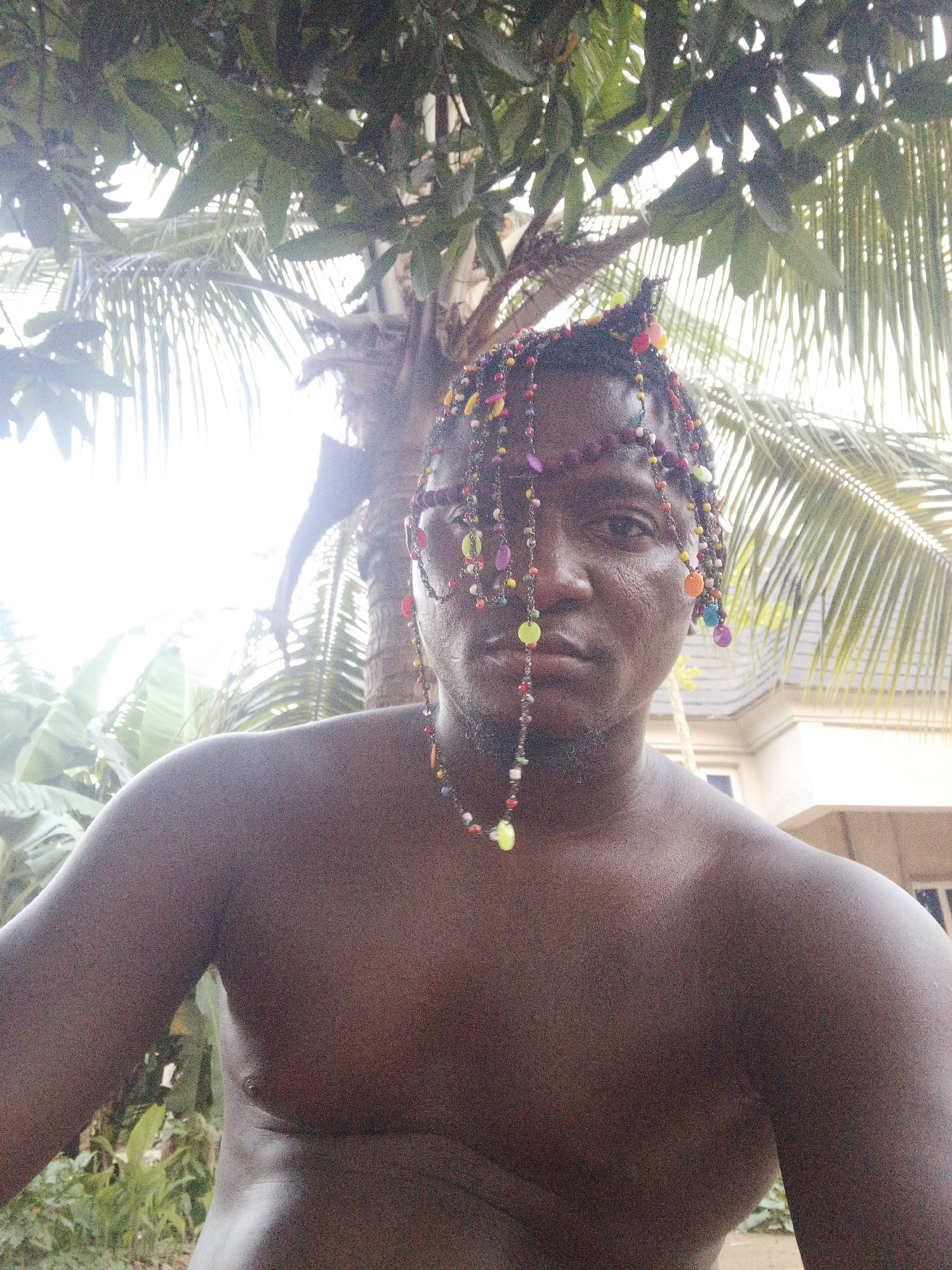 An outdoor portrait shot in Akwanga, Nigeria, features a dark-skinned man with an intense, direct gaze towards the camera. He is shirtless, revealing his bare chest, shoulders, and upper torso. His hair is intricately adorned with numerous colorful beads of various shapes and sizes, forming strands that drape across his forehead, down the sides of his face, and around his neck. The beads are predominantly red, yellow, green, blue, purple, orange, and white. Some strands partially obscure his left eye and cheek.
The man's skin appears slightly glistening, possibly from sweat, reflecting the bright natural light. He maintains a serious, neutral expression.
The background is filled with lush green foliage, including large leaves and distinct palm fronds, suggesting a verdant, possibly tropical environment. The strong light from behind the subject creates some overexposed areas in the background, indicating a sunny day, likely midday or early afternoon. A light-colored building is partially visible in the distant background to the right, suggesting a residential or developed area within the natural setting. No visible text is present in the image.