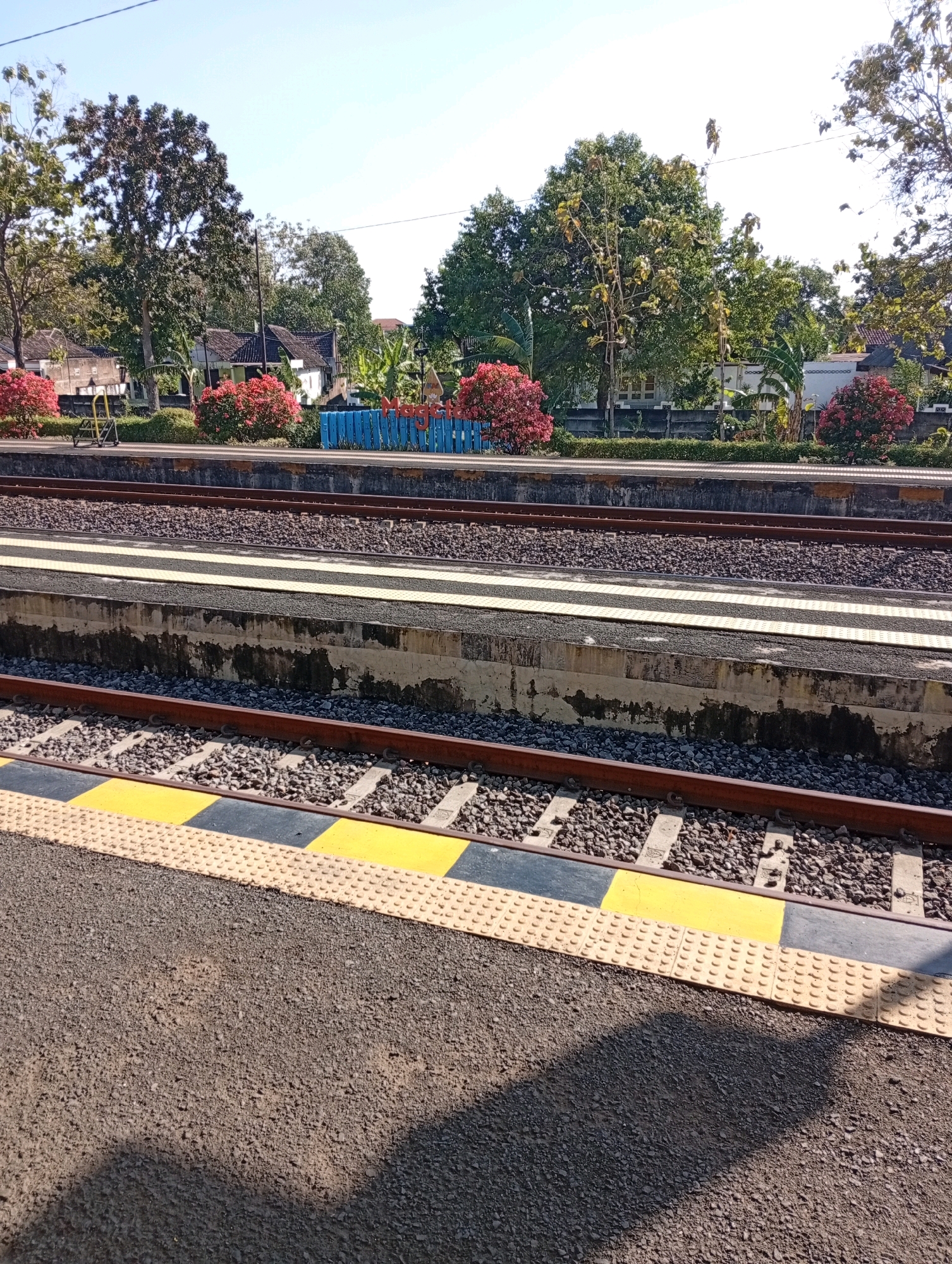 This image captures a view of the Tebon train station in Tebon, Indonesia, on a clear, sunny day. The foreground shows the edge of a train platform with a tactile paving surface consisting of raised circular bumps, bordered by yellow and black safety strips. Beyond this, a railway track is visible, laid on a ballast of crushed stone and supported by concrete sleepers.
Further back, another platform and track are visible. The platforms are constructed with concrete edges. A blue fence with red lettering, potentially indicating the station's name or a welcome message, runs along the back of the platforms. Behind the fence, there is a line of vibrant red flowering bushes and lush green trees. In the distance, residential buildings with dark tiled roofs are nestled amongst the greenery, suggesting a semi-rural or suburban setting. The sky is a bright, clear blue. There are no people or animals visible in the image. The scene is static and appears to be an architectural and environmental overview of the station.