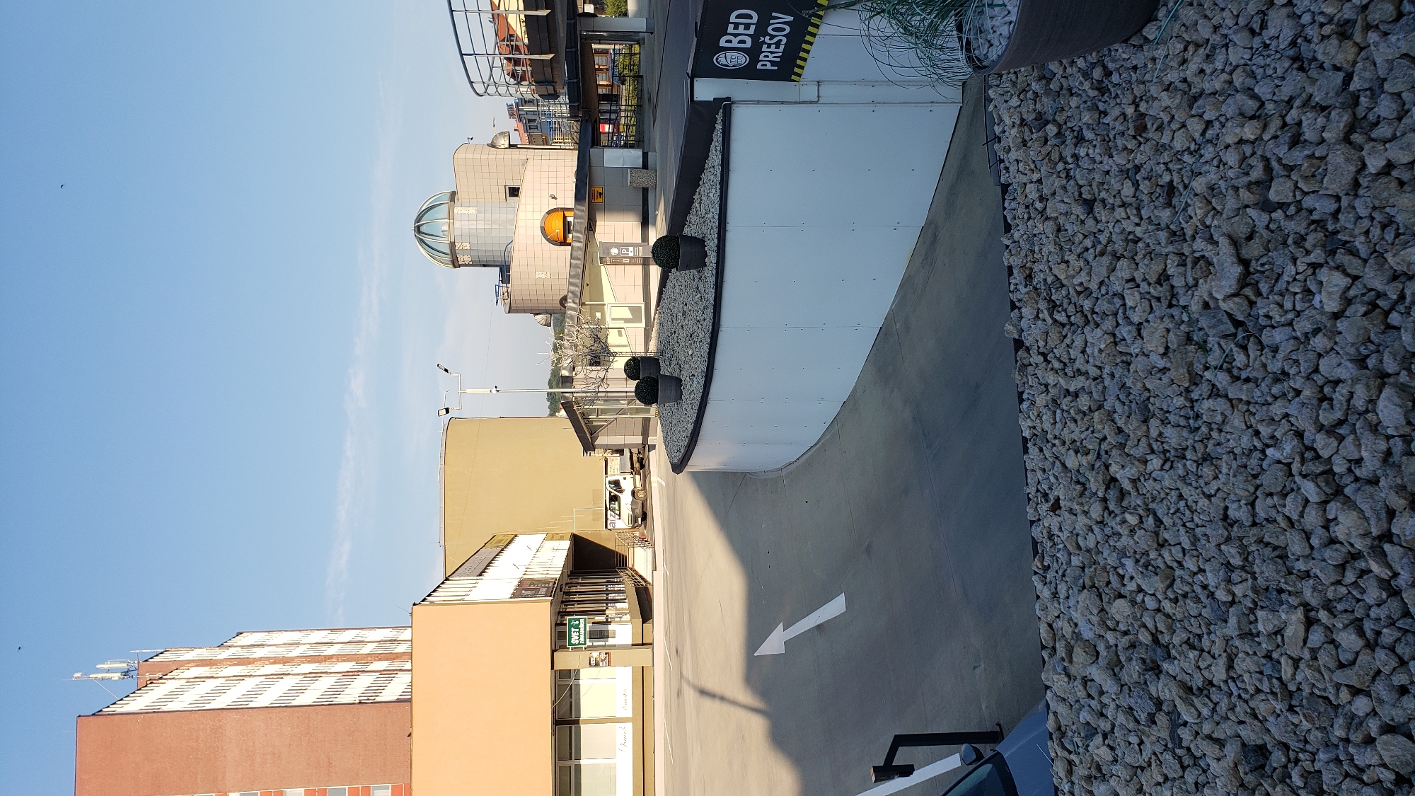 The image depicts an outdoor scene in the city of Prešov, Slovakia, featuring modern architecture and urban elements. In the foreground, a paved area with a white arrow indicating direction leads into a curved concrete structure with a white facade. This structure is adorned with small decorative plants in pots and covered in gravel. To the right, a sign with a black and yellow border reads "BED PREŠOV".
Behind this structure, a multi-story building with a distinctive domed roof and glass elements is visible. Adjacent to it, a beige building with multiple windows and a signage that reads "SVET" and "Dance!" is present, suggesting commercial or retail establishments. A white van is parked near the beige building.
In the background, a tall brick building with many windows stands prominently, likely an apartment complex or office building. The sky is clear and blue, indicating fair weather. The overall impression is one of a modern urban environment, possibly a commercial district or a civic center within Prešov.