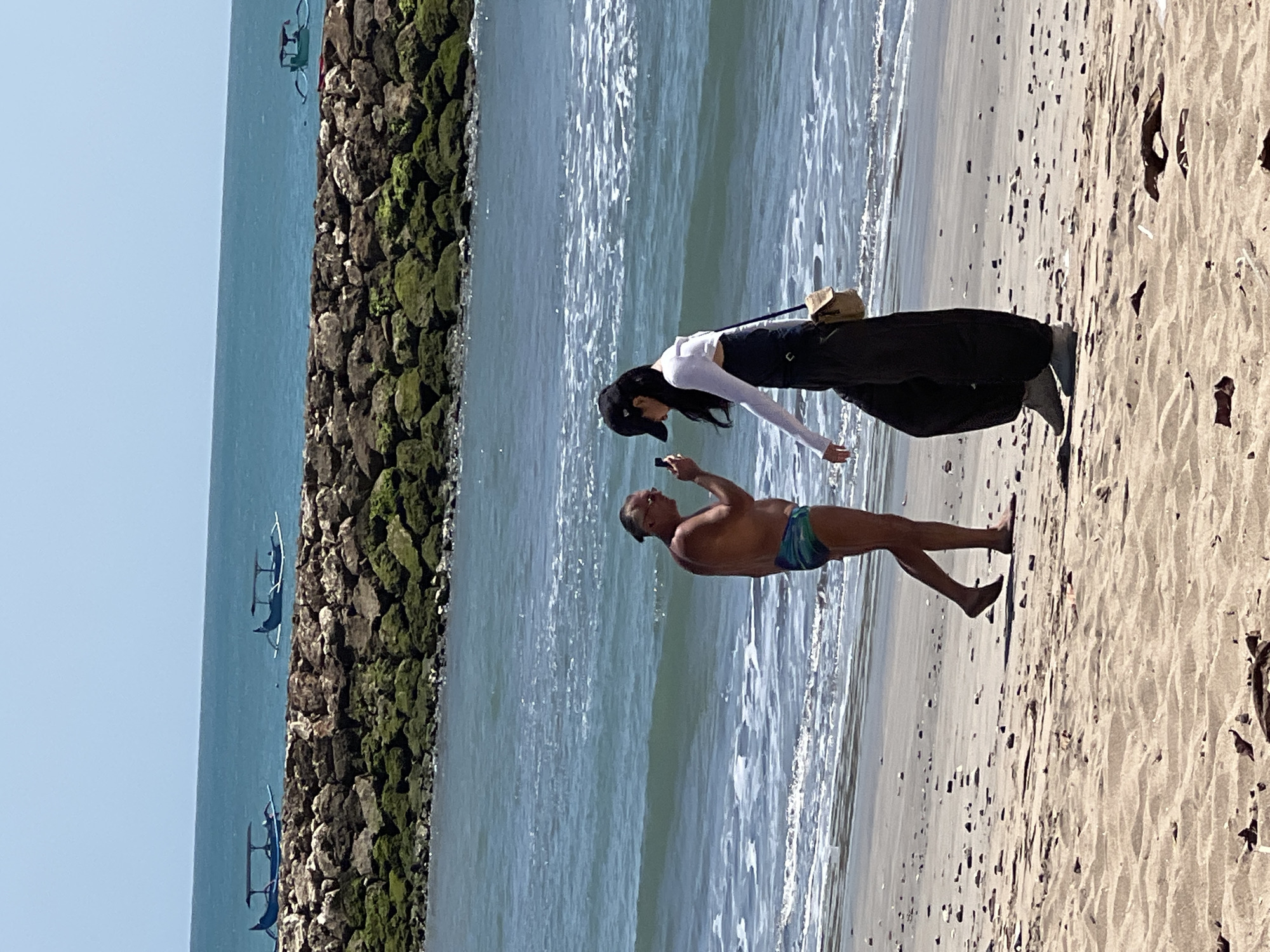 On a sandy beach in Kuta, Bali, Indonesia, two individuals are engaged in an interaction. A man, wearing swim trunks, stands near the water's edge, holding a phone. Facing him is a woman, dressed in dark pants and a white long-sleeved top, with a black cap on her head. She appears to be looking at the man's phone, possibly taking a picture or video. The ocean, with gentle waves and white foam, is behind them. In the distance, a stone breakwater extends into the sea, with several traditional Balinese fishing boats, known as jukung, visible on the water. The sky is clear and blue. The overall scene suggests a casual moment on a sunny day at the beach.