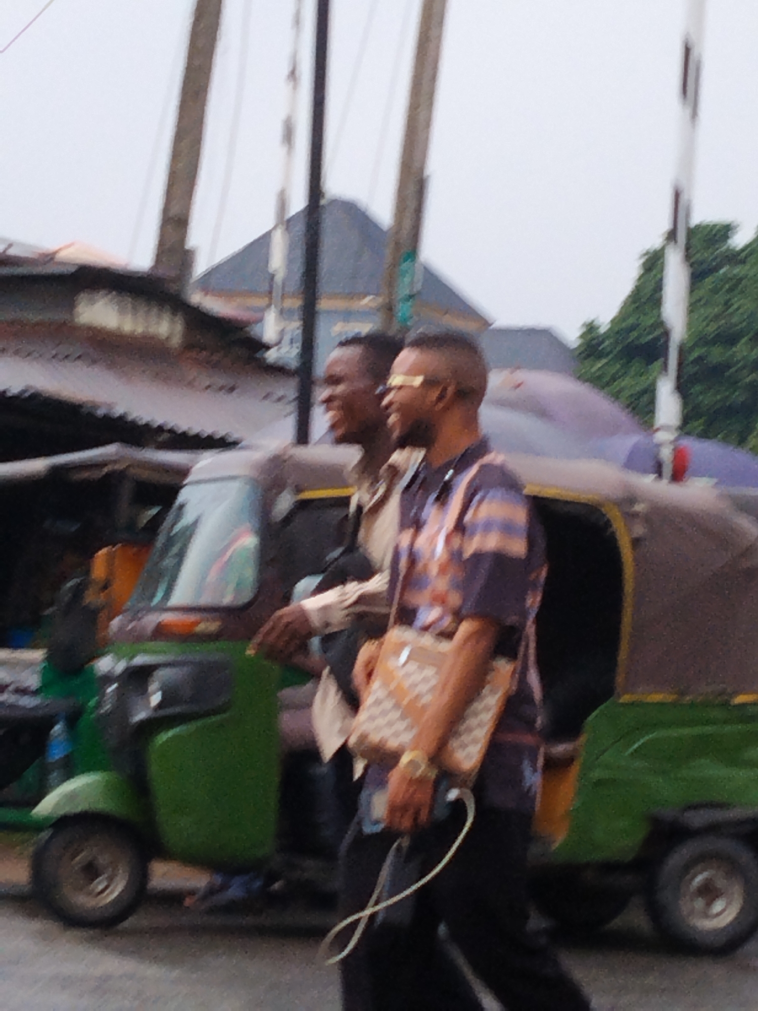 Two men are walking along a street in Umuode, Nigeria. One man is wearing a light tan shirt and dark pants; the second man wears a dark short-sleeved shirt with orange and blue horizontal stripes, dark pants, a beige messenger bag, and yellow sunglasses. They are smiling.
Parked beside them is a three-wheeled motorized vehicle, painted green and brown. The environment is partially urban, with buildings in the background and utility poles overhead.
