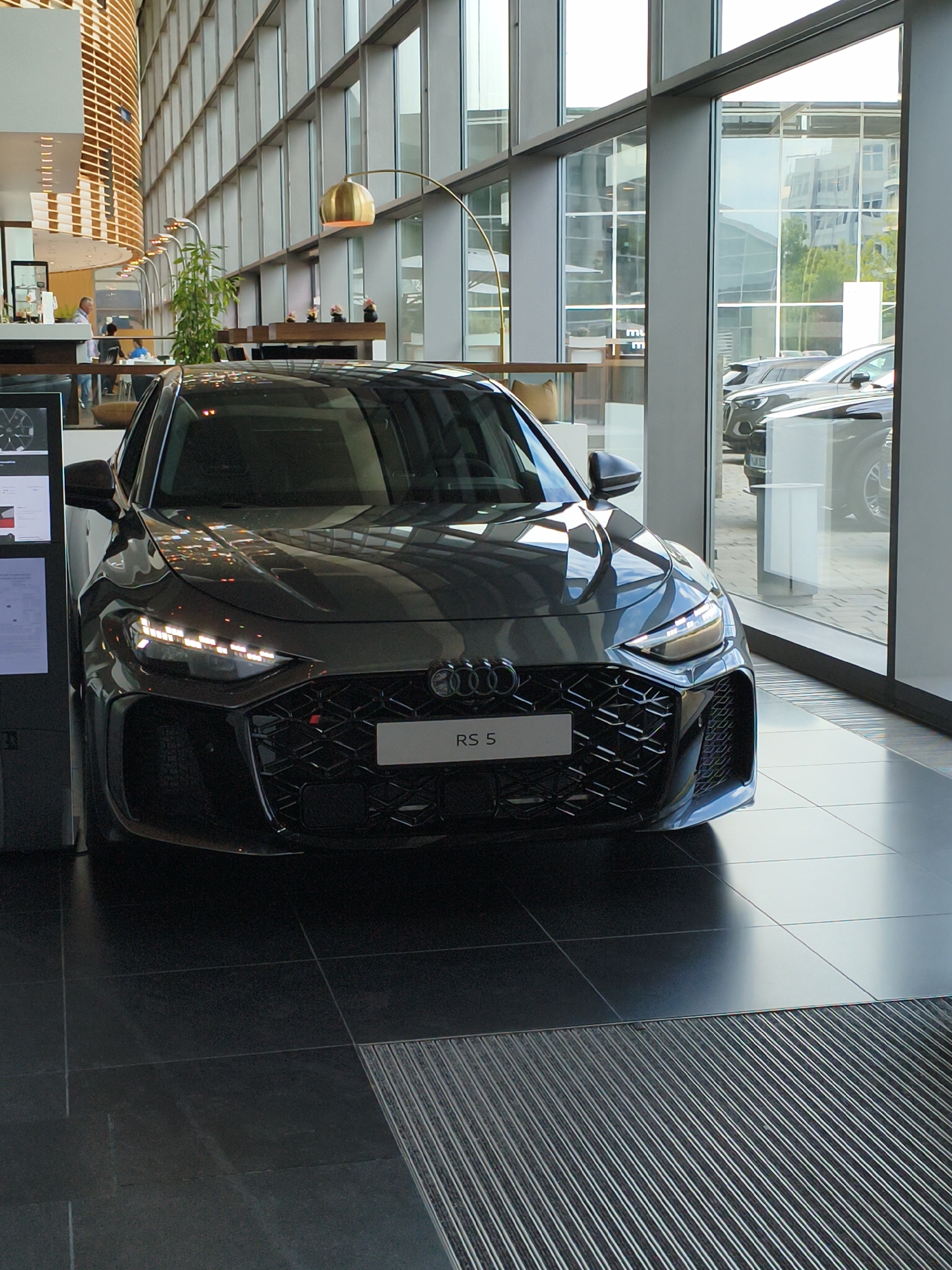 The image shows the front view of a dark gray Audi RS 5 parked indoors, likely in a showroom or exhibition space. The car is positioned at an angle, facing slightly to the right of the camera. Its headlights are illuminated, casting a bright glow. The grille features a honeycomb pattern, and a white license plate with "RS 5" is visible in the center.
Behind the car, a large, modern interior is visible, characterized by a high ceiling and expansive windows that reflect the surrounding environment. The interior architecture includes curved wooden slats on the left wall and vertical window frames. In the background, through the windows, other cars, possibly other Audi models, can be seen parked outside or in an adjacent area. Some people are visible in the background, appearing to be employees or visitors in the space, interacting with each other or observing the vehicles. A tall golden floor lamp with a curved arm is positioned to the left of the car, adding a touch of ambiance to the scene. The floor is tiled with large, dark gray squares, with a ribbed mat in the foreground. The lighting suggests it's daytime, with natural light filtering in through the large windows. The environment appears clean, modern, and well-maintained, typical of a high-end car dealership or corporate space.
The image does not provide any specific information about the city of Ingolstadt, Germany.