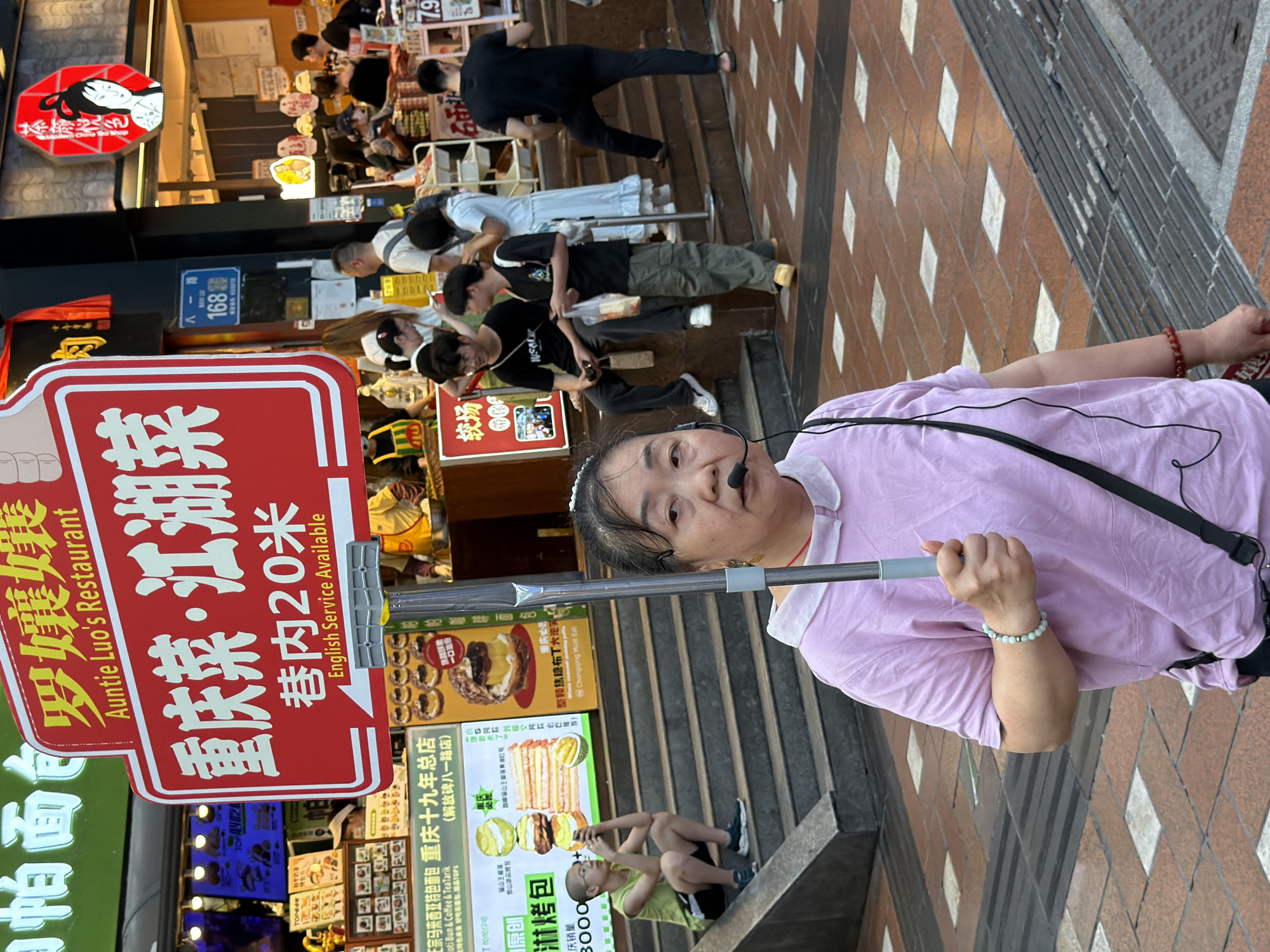 This media file shows a street scene in Yuzhong District, China. The foreground features a woman wearing a pink shirt, a headset, and a bracelet, holding a selfie stick. Behind her, a red sign with Chinese text and an English translation reads "Auntie Luo's Restaurant" and "Chongqing Dish · Jianghu Dish" with "20 meters inside" and "English Service Available". Further in the background, several people are seen walking or standing, with shops and advertisements lining the street. One advertisement displays pictures of food items, including buns and cakes, with Chinese text. The scene appears to be outdoors during daylight, possibly in a busy commercial area given the shops and pedestrian traffic. The woman's expression is neutral, and the overall mood of the scene is that of a typical day in a bustling urban environment.