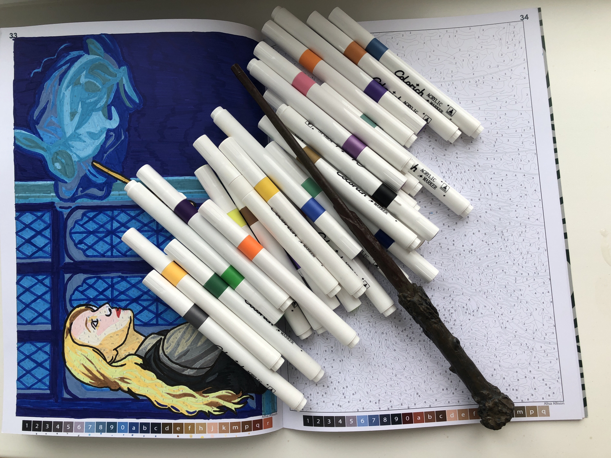 The image depicts an open book, likely a coloring book, on a white surface. The book is opened to two pages, numbered 33 and 34. Page 33 features a partially colored image of a woman with blonde hair looking upwards, a window behind her, and a blue, ghostly animal figure above the window. Page 34 appears to be a "color-by-number" image, still uncolored, with various small numbers and letters indicating which color to use in each section.
Scattered across the book are numerous white acrylic markers with colored bands around their caps. The colors visible include orange, pink, purple, green, and blue. A brown, textured wand, resembling a magic wand, is also placed on top of the book and the markers. The markers are labeled "Colorich ACRYLIC MARKER." The bottom of page 33 shows the colors needed for that page, labeled "1 2 3 4 5 6 7 8 9 0 a b c d e f h j k m p q r". The bottom of page 34 shows the colors needed for that page, labeled "1 2 3 4 5 6 7 8 9 0 a b c d e f m p q". The book is credited to "Alisa Allison" on both pages.
There is no indication of location in the city of Noviy, Russia.