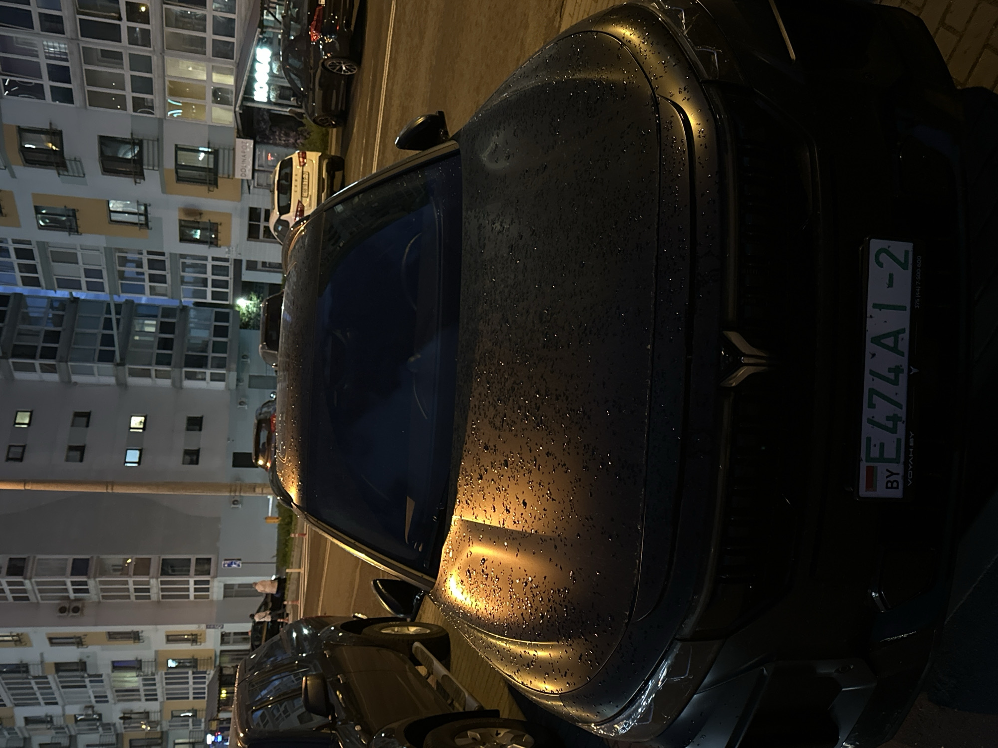 The image shows a dark-colored Voyah Free car parked in an urban setting, likely in Minsk, Belarus, based on the "BY" license plate and Cyrillic text. The car's license plate reads "E474AI-2". It appears to be night or dusk, as indicated by the lighting and the illuminated windows of a multi-story building in the background. The car is wet, likely due to rain. Other cars are parked nearby, suggesting a parking lot or a street with parked vehicles.