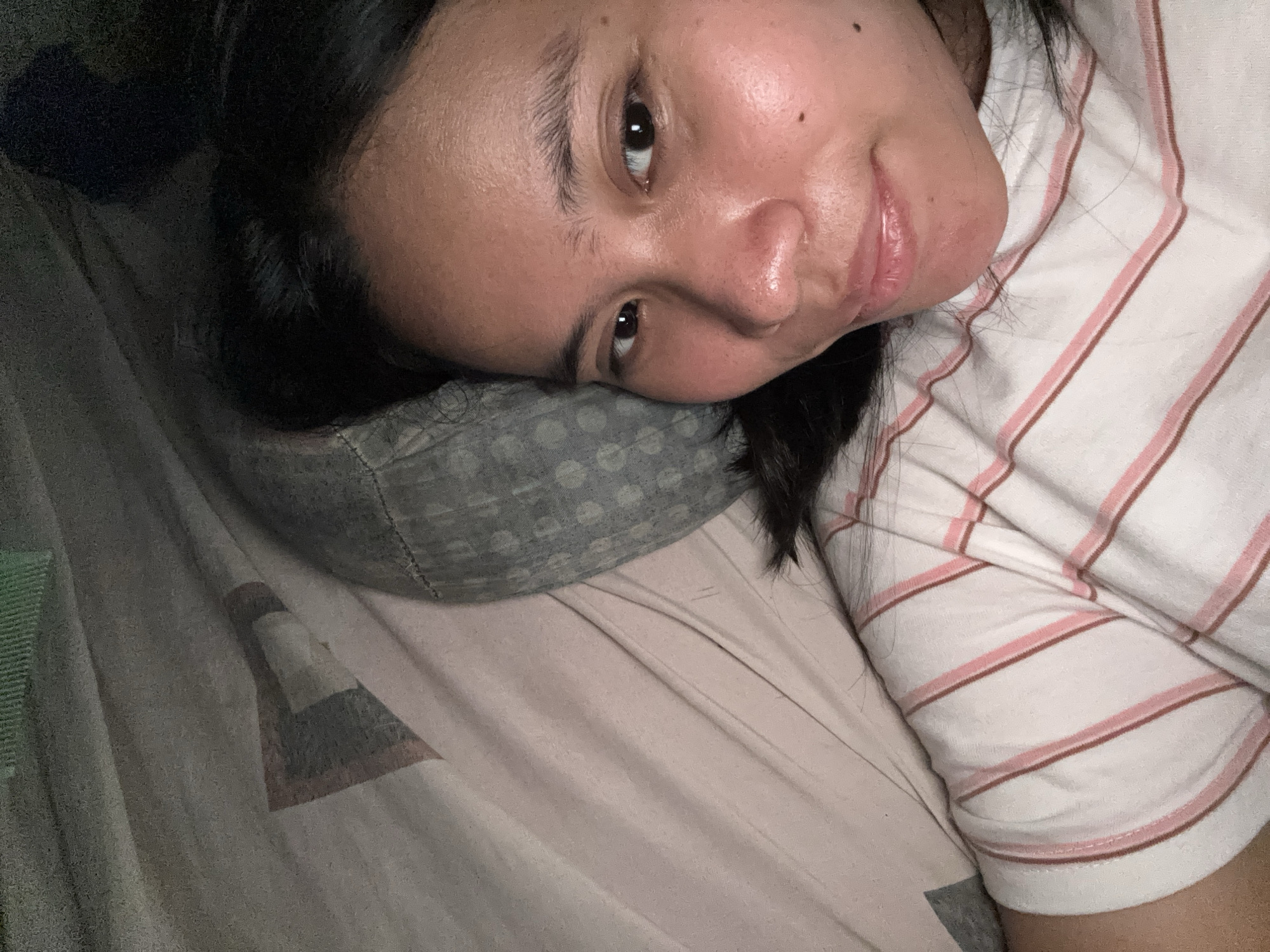 The image is a close-up, low-angle selfie of a young woman lying on her side on a bed. She has dark hair, brown eyes, and light brown skin. She is wearing a white t-shirt with pink and brown stripes. The background consists of bedding with a striped pattern. The image is slightly blurry and appears to have been taken in low light conditions, possibly indoors in a bedroom. There is no discernible location in the city of East Jakarta, Indonesia, within the image itself.