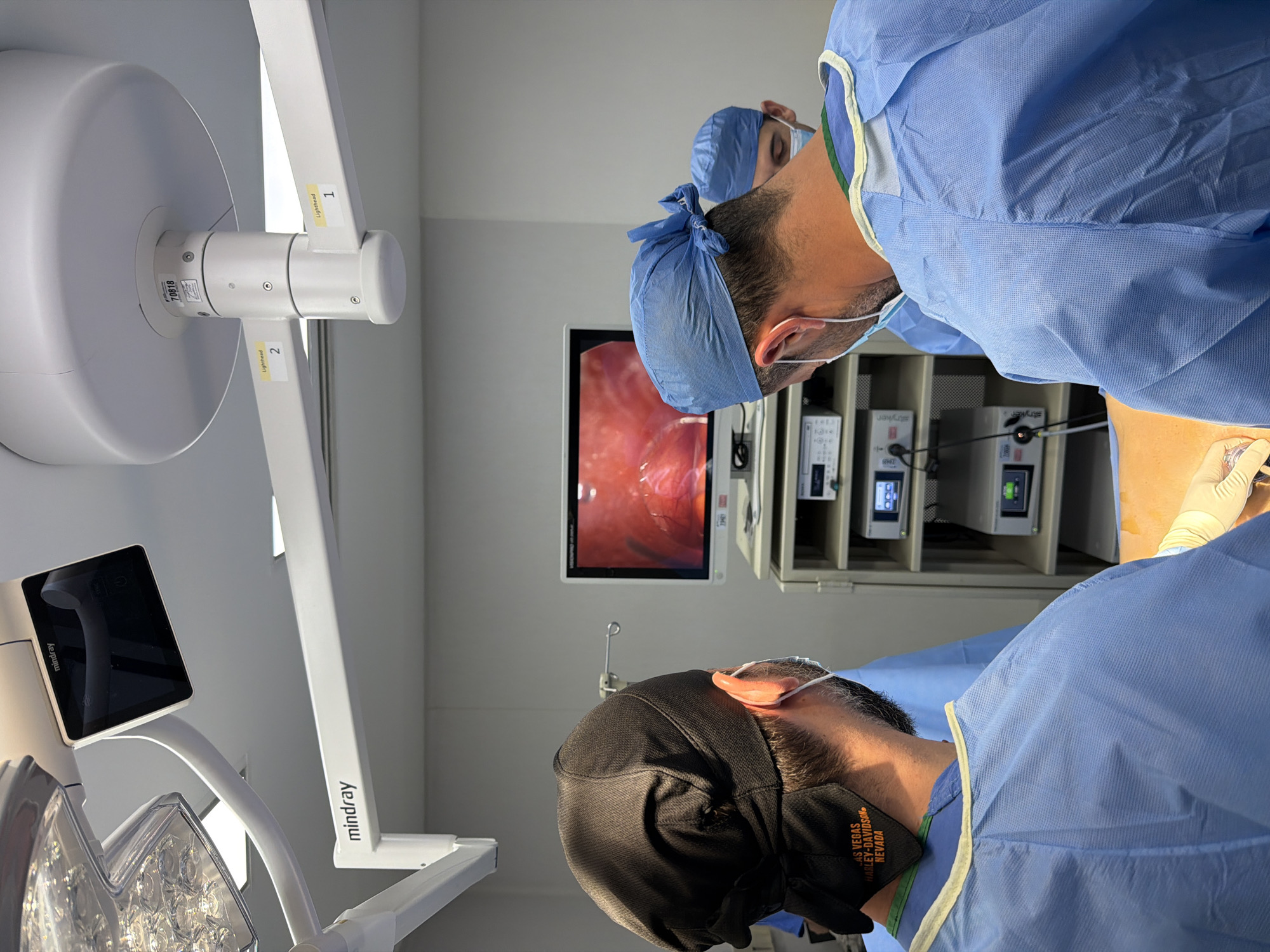 The image shows two individuals, presumed to be medical professionals, participating in what appears to be a surgical procedure. Both individuals are wearing surgical attire, including gowns, caps, and masks. One individual is wearing a black mask with "LAS VEGAS HARLEY-DAVIDSON NEVADA" printed on it. They are both focused on a monitor displaying a live endoscopic feed. Medical equipment, including a "mindray" branded surgical light and "Stryker" branded medical devices, are visible in the background. The image doesn't feature Puebla, Mexico. It is in a surgical room.