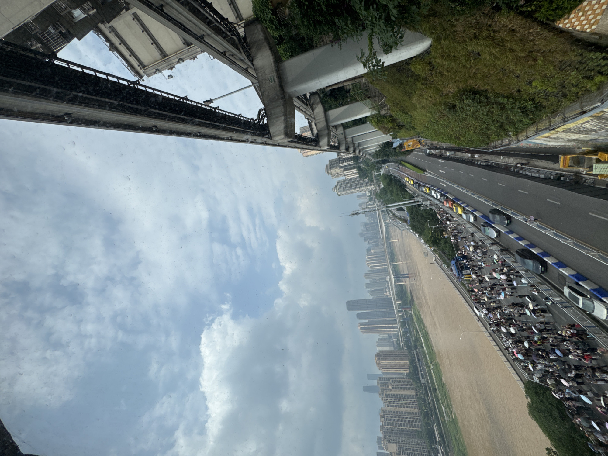 The image shows an elevated view of a scene in Yuzhong District, China. A large group of people with umbrellas is visible along the riverfront. The buildings in the background suggest a dense urban environment. A highway runs parallel to the water. Above, a monorail structure and its supporting pillars are prominent, with the sky visible through gaps in the structure.