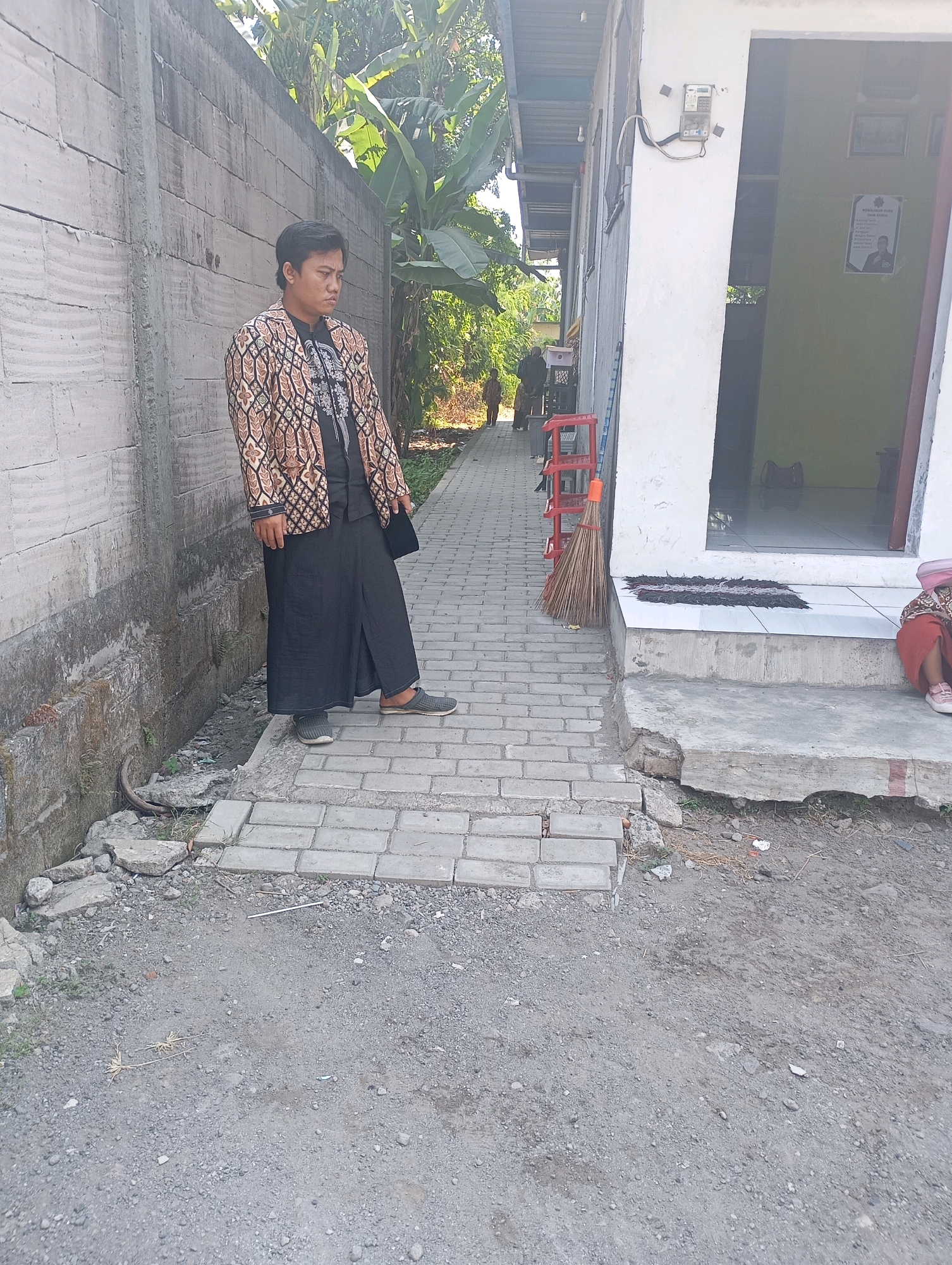 In Ketegan, Indonesia, a young person stands on a tiled walkway next to a concrete wall. They are wearing a patterned jacket over a dark shirt and a long black skirt. The walkway is bordered by a rough, gravelly ground. To the right of the walkway, there is a building with a white facade and an open doorway leading into a room with green walls. A broom leans against the building. A red plastic chair is visible further down the walkway. In the background, lush green foliage and trees are present. The scene appears to be outdoors on a sunny day.