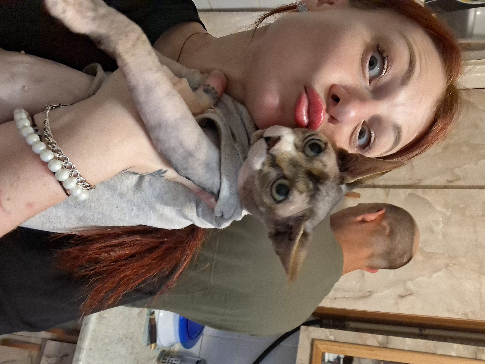 A medium shot shows a woman with red hair pouting her lips and holding a Sphynx cat. The woman is on the left side of the frame, looking at the camera with wide eyes. The cat, wearing a grey sweater, is held in her arms, and its head is close to the woman's face. In the background, a bald man in a green shirt is visible, his back to the camera, facing a counter. The setting appears to be a kitchen, with cabinets and a countertop visible. The lighting is artificial. The overall impression is a casual, selfie-style photograph.