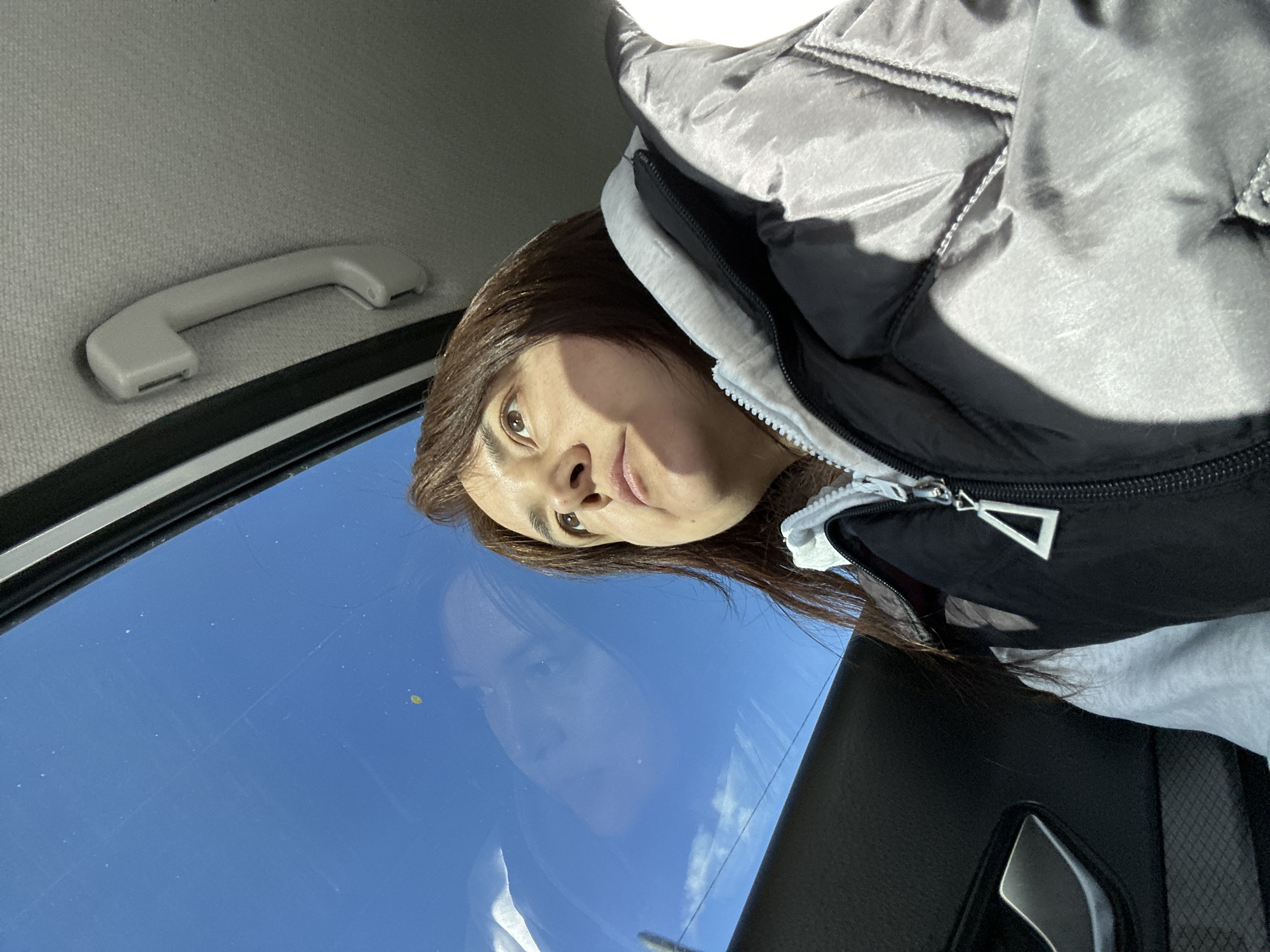 The image shows a person inside a car. The person is wearing a jacket with a black and white color scheme. The car's interior includes a grab handle, a window showing a clear blue sky, and a black door panel. The person appears to be looking upwards. The location is Unknown, Unknown.