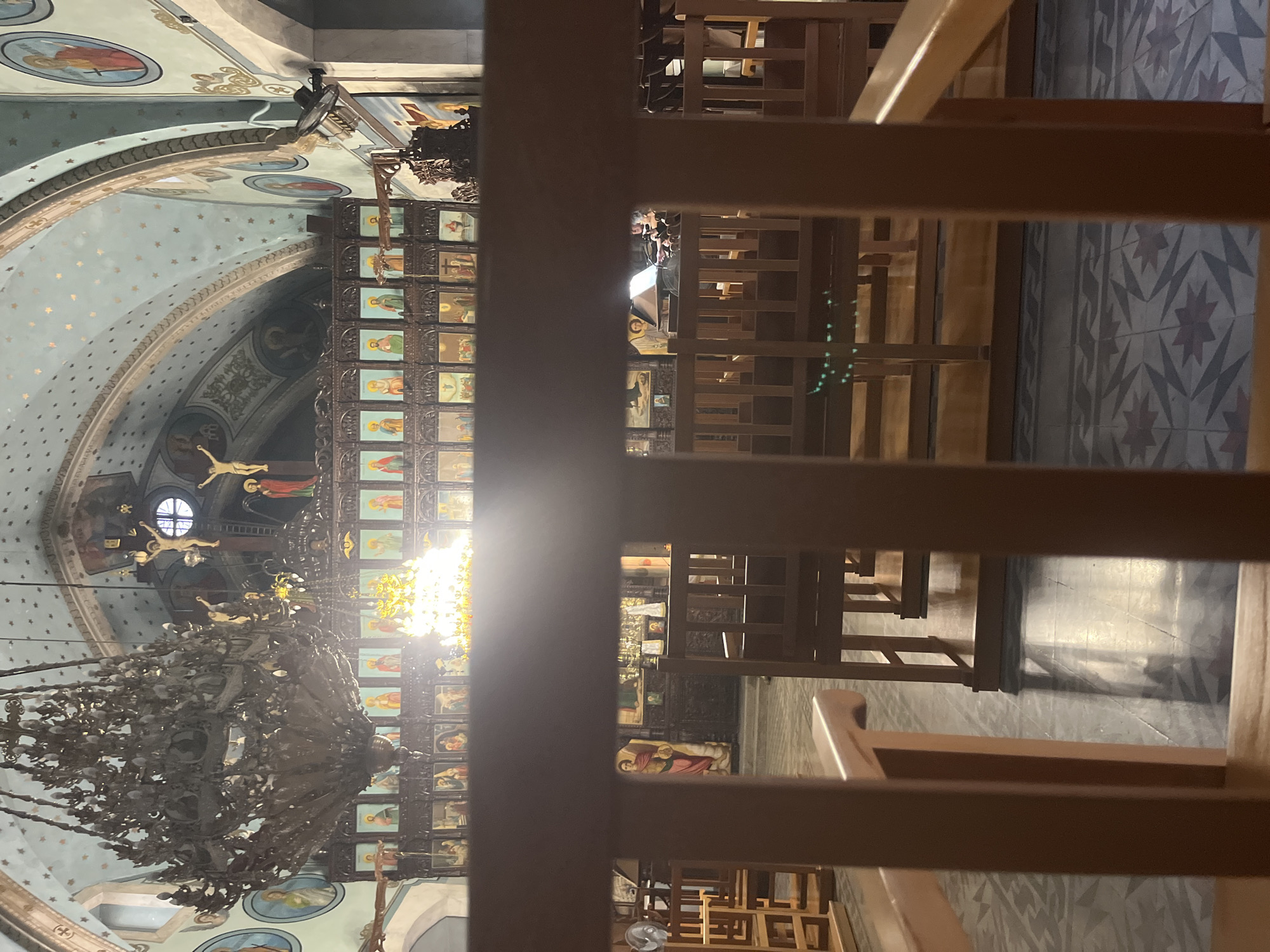 The image depicts the interior of the Holy Church of Panagia Iamatiki in Lofou, Cyprus. The foreground shows the backs of wooden pews, suggesting the viewer is seated. Beyond the pews, an iconostasis, a wall of icons, spans the width of the church, adorned with religious imagery. Above the iconostasis, a large, ornate chandelier hangs prominently, casting a warm glow, and a smaller, brightly lit chandelier is visible further in. The ceiling is decorated with a light blue hue and sprinkled with golden stars. Religious iconography, including depictions of Jesus Christ on the cross, are visible throughout the space. The floor features a patterned tile design. The overall atmosphere is solemn and reverent, characteristic of a place of worship.