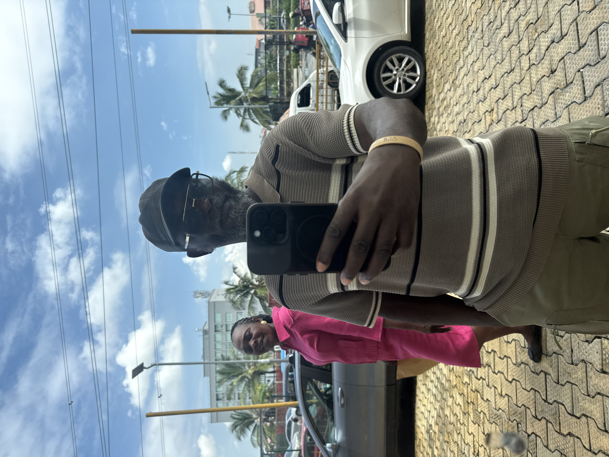 This is a vertical, eye-level, full-body selfie of a Black man, taken outdoors on a sunny day. He is wearing a brown and white striped sweater, khaki pants, glasses, and a dark baseball cap. On his left wrist, he wears a yellow bracelet. He is holding a black smartphone, presumably to take the photo, and is looking at the camera.
To his left and slightly behind him, a Black woman is visible. She is wearing a bright pink jacket and dark pants. She is looking towards the camera and smiling.
The background suggests an urban environment in Itirin, Nigeria. Palm trees are visible, along with buildings and a white car. The ground is paved with interlocking bricks. The sky is blue with scattered white clouds, indicating fair weather. There are power lines running horizontally across the upper part of the image. A yellow pole, possibly part of a traffic light or street sign, is visible in the distance. The overall mood appears casual and pleasant.