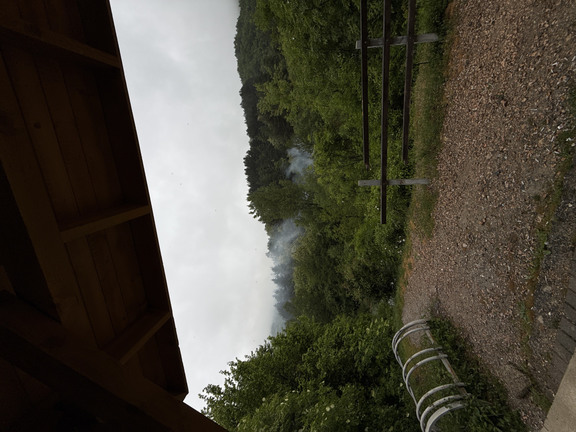 The image shows a view from under a wooden structure, possibly a shelter or gazebo, with the landscape visible beyond. A path composed of small stones or gravel is visible to the right. A simple metal fence or barrier is present near the path. In the distance, there is a forest area with trees. Smoke can be seen rising from within the forest. The sky is overcast. Based on the provided information, the location is Ljevoša - Lëvoshë, Kosovo.