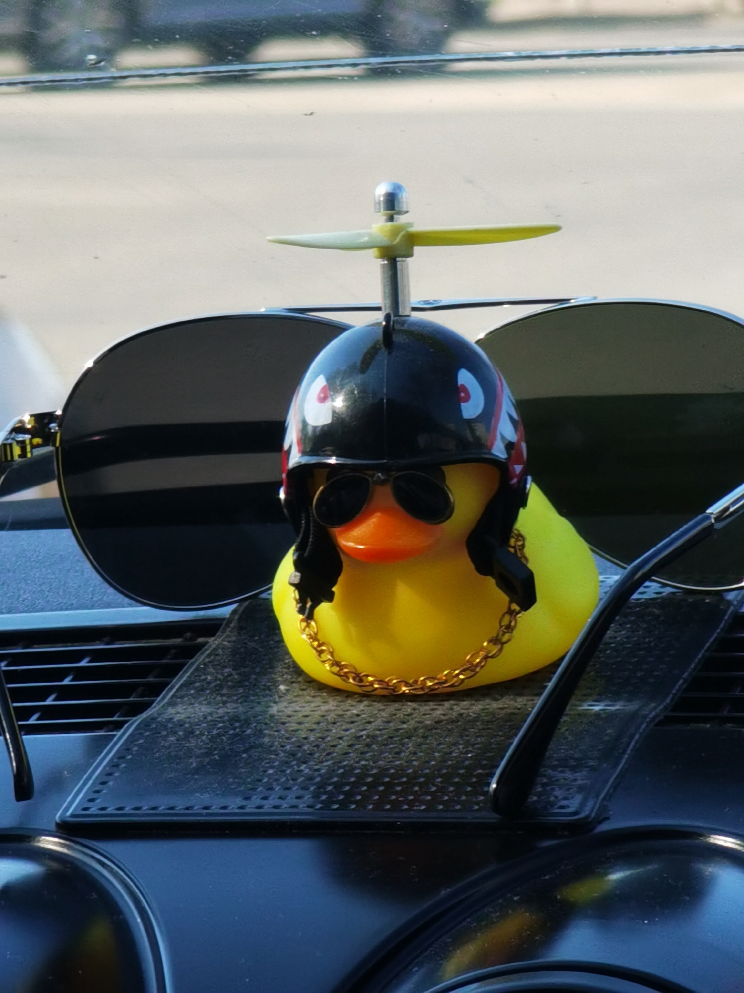A yellow rubber duck with an orange bill rests on a black textured surface, likely a car dashboard. The duck wears a black helmet with cartoonish red and white accents and a small propeller on top. It also sports sunglasses and a gold chain around its neck. Behind the duck, a pair of sunglasses with dark lenses are positioned. The background shows a blurred street scene, possibly in Chaikovsky, Russia, though specific landmarks are indistinguishable.