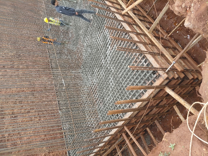 The scene depicts an active construction site in Fdalate, Morocco, focused on the foundation and basement structure of a building. An excavated pit, with walls of reddish-brown soil, contains the ongoing work.
In the center, a large, rectangular grid of steel reinforcement bars (rebar) forms the base for a concrete slab. This grid is partially filled with wet concrete, suggesting a recent pour. Vertical rebar extends upwards from this slab, intended for structural walls. Wooden formwork, constructed from planks and timber supports, lines the perimeter of the pit, encasing the vertical rebar along the edges of the concrete slab.
Four individuals, identified as construction workers, are present within the pit. One male worker, dressed in a dark long-sleeved shirt and grey trousers, walks on the rebar grid and partially set concrete in the mid-ground. In the background, two other male workers are visible. Both wear yellow hard hats and high-visibility vests. One of these workers appears to be operating a vibrating tool or hose, directed at the concrete near the excavated wall. A fourth worker is partially visible on the far right, also within the pit.
The environment is an open-air excavation under natural daylight. White electrical wiring is visible on the reddish-brown soil in the bottom-left foreground, outside the main pit.