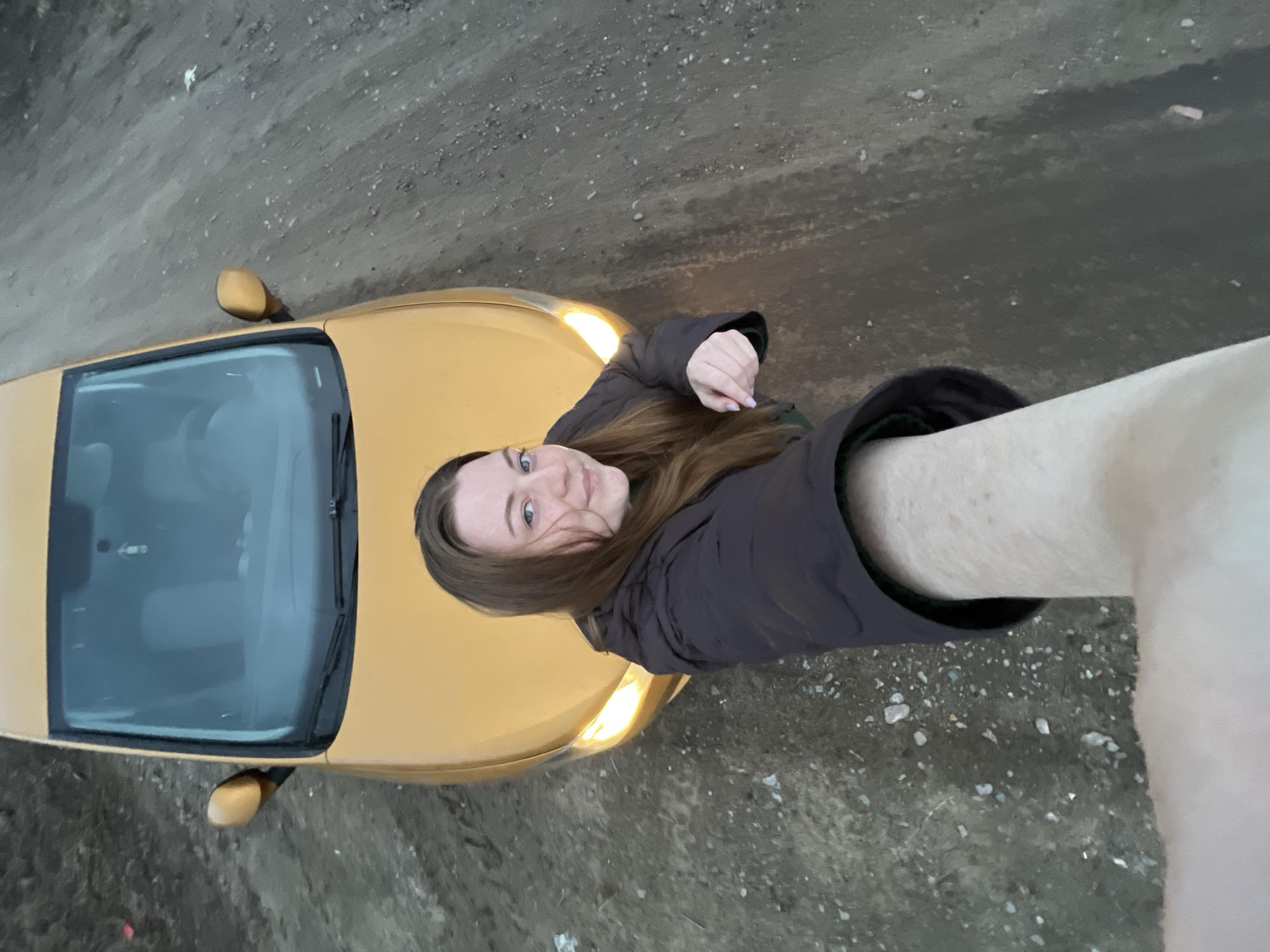 A selfie photograph taken outdoors, likely in Раздолье, Russia, shows a young woman with brown hair wearing a brown jacket. She is positioned to the right of a yellow car, and the photograph is taken from an elevated angle. The car's headlights are on. The ground appears to be a dirt or gravel road. The sky is overcast.