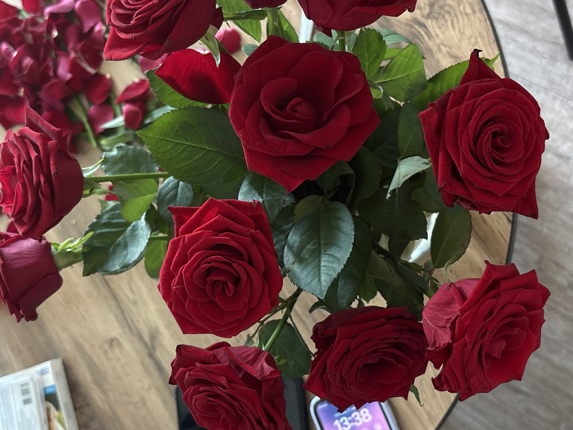 The image shows a bouquet of red roses arranged on a wooden table. The roses are in various stages of bloom, with some fully open and others still in bud. Their petals are a deep, rich red, and their leaves are a dark green. A smartphone is visible on the table, displaying the time "13:38". In the background, a stack of white boxes or cases can be seen. The scene appears to be indoors, likely on a table, and there are no people or animals present in the image. The overall impression is one of elegance and beauty, with the vibrant red of the roses contrasting with the natural tones of the wooden table. The image does not provide any specific visual cues that would allow for an accurate identification of its location within Rostov-on-Don, Russia.