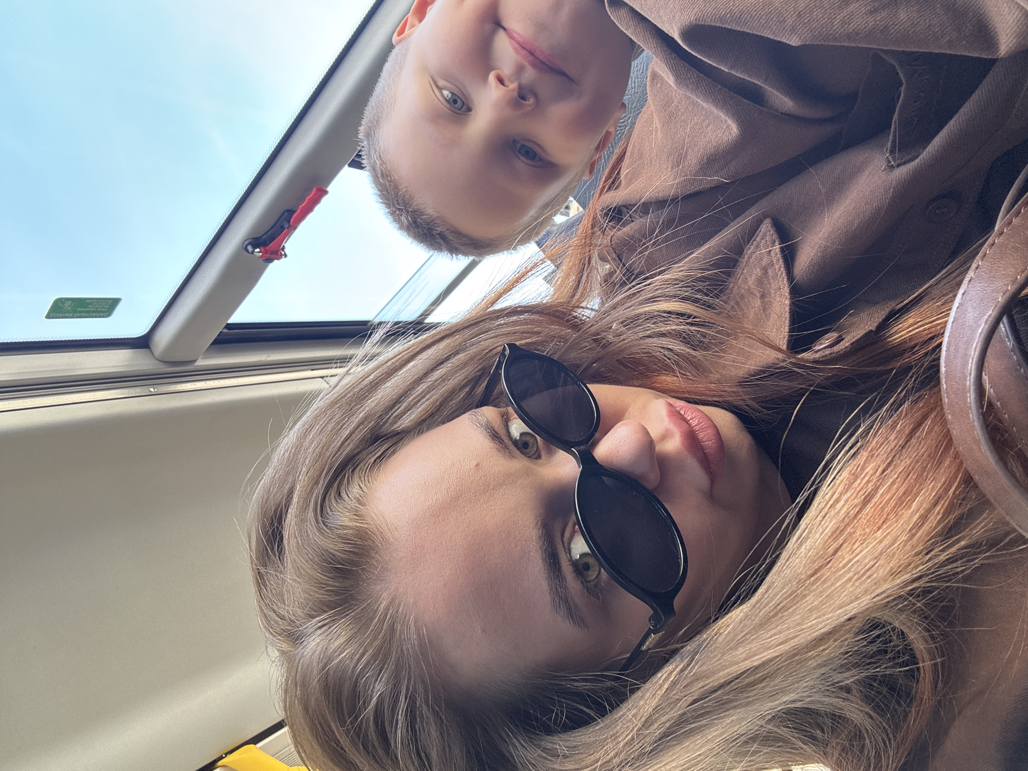 Two people are in the frame. One is an adult woman wearing black sunglasses and a brown jacket. She is positioned in the foreground, facing towards the camera. The other person appears to be a child with short hair, also wearing a brown shirt or jacket, positioned above and slightly behind the woman. They both appear to be inside a vehicle with a sunroof. Visible through the sunroof is a section of blue sky. Part of the car's interior trim is visible near the sunroof. The location cannot be verified as Chaikovsky, Russia, based solely on the image.