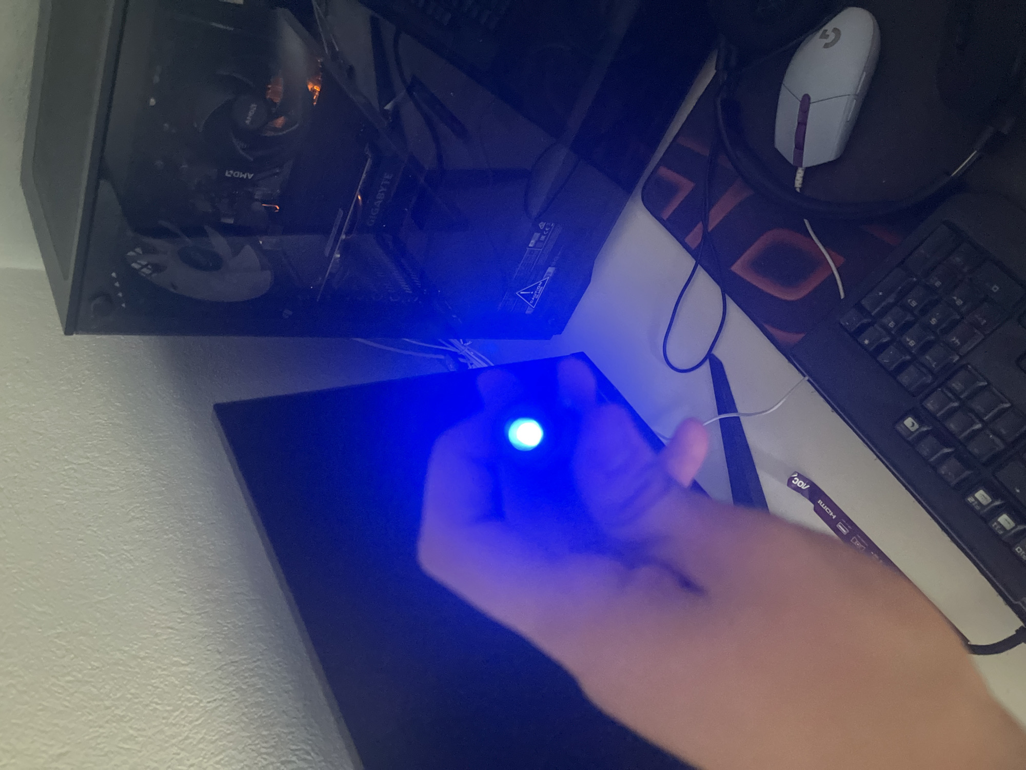 The image shows a hand holding a small blue LED light in front of a computer setup. A computer tower with visible internal components and lighting is on the right. To the left, a black monitor displays a blue light. A keyboard and mousepad are visible in the foreground. The environment appears to be an indoor desk area, likely a home or office, with a textured wall in the background. There is no information or visual evidence in the image to indicate a location in Pokrovnik, Bulgaria.