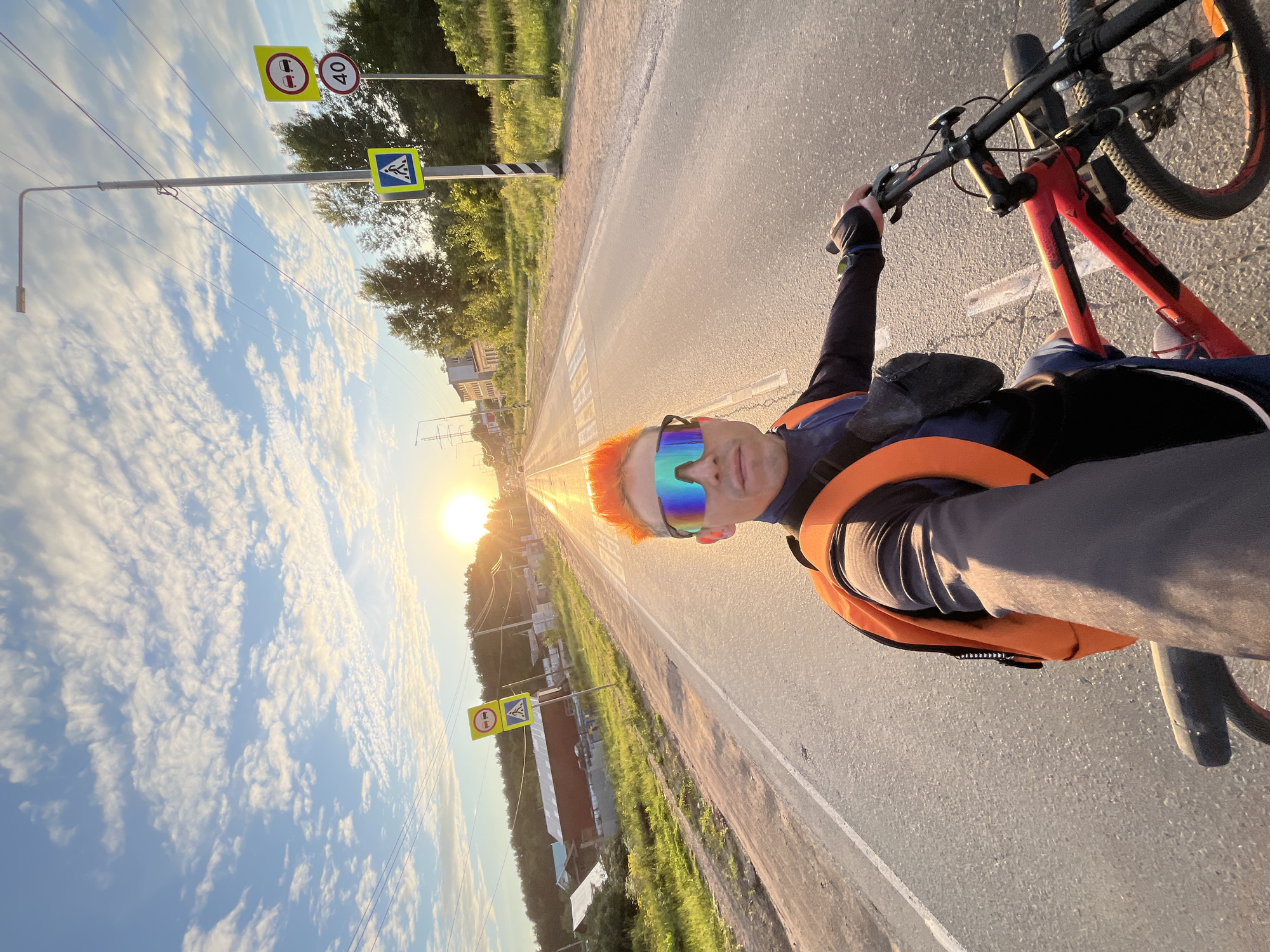 An outdoor, eye-level selfie captured by a male cyclist on a paved asphalt road at sunset in Chaikovsky, Russia. The man, positioned on a red mountain bicycle, occupies the lower right quadrant of the frame, holding the camera at arm's length. He has bright orange hair and wears mirrored, iridescent sunglasses, a black long-sleeved top, an orange backpack, and a black fanny pack across his chest. His right hand rests on the bicycle's handlebar.
The road, illuminated by the low sun, stretches straight into the background towards the horizon, where the sun appears as a bright, circular disc. White solid and dashed lines are visible on the road surface, with a pedestrian crossing marked by white dashed lines further in the distance.
On the right side of the road, a tall, grey street light pole stands prominently, accompanied by multiple power lines running horizontally across the sky. Below the street light, a cluster of Russian road signs is visible: a yellow square pedestrian crossing sign (5.19.1), a round white "No Overtaking" sign (3.20), and a round white "Speed Limit 40" sign (3.24). Adjacent to the road on both sides is green vegetation, including trees and bushes.
On the left side of the road, in the mid-ground, multiple utility poles with wires are present, and beyond them, several low-rise buildings, including one with a reddish-brown roof, are visible amidst the greenery. The sky above is a clear blue with scattered white altocumulus clouds, reflecting the warm hues of the setting sun.