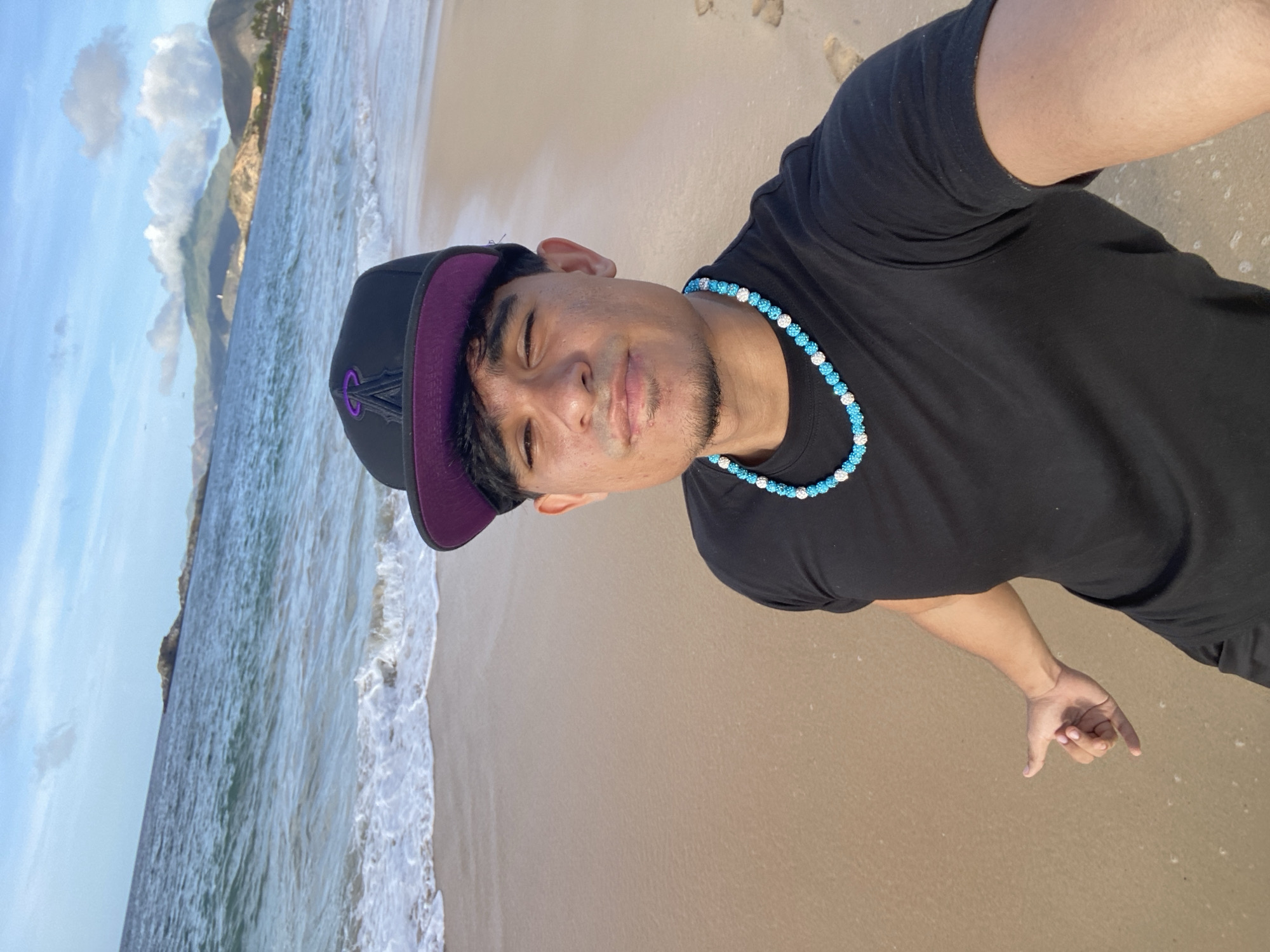 A young man wearing a black t-shirt, a black baseball cap with a purple brim and logo, and a blue and white beaded necklace is taking a selfie on a sandy beach. He is standing with the ocean and a coastline with mountains in the background. He is making a "shaka" hand gesture with his left hand. The scene appears to be a bright, sunny day.