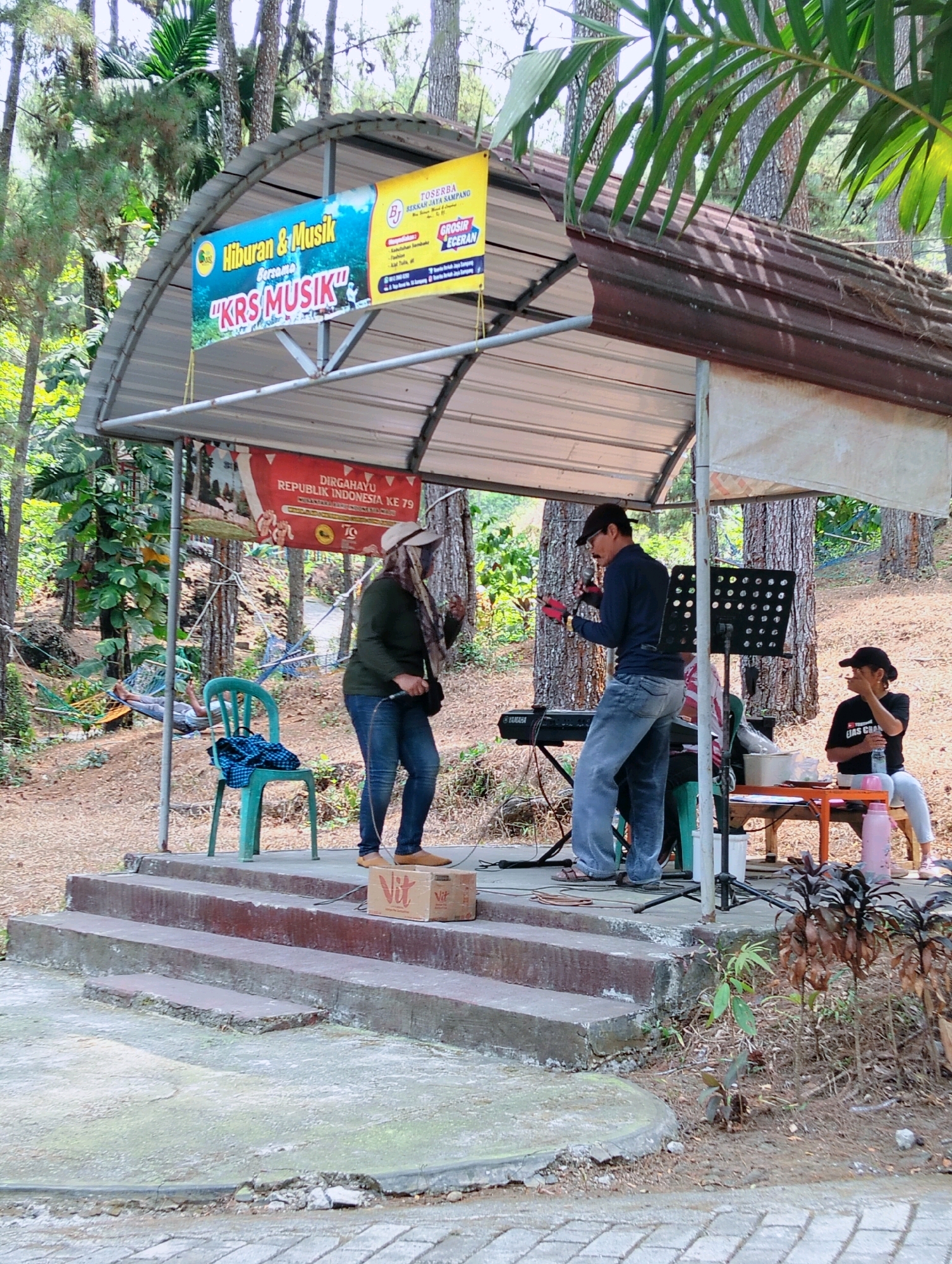 The image depicts an outdoor stage set up in a wooded area, likely a park or recreational space. Two individuals are performing on stage. One, a person wearing a hat and a dark long-sleeved shirt, is singing into a microphone, holding it in their right hand. They are dressed in jeans. The other performer, a woman wearing a hat and a dark long-sleeved shirt, is also holding a microphone and appears to be singing or speaking. She is also wearing jeans.
In the background, a keyboard is visible, along with a music stand. A green plastic chair with a dark checkered bag on it is placed to the left of the stage. To the right, a person wearing a black t-shirt and a cap is seated on a bench behind a small orange table, holding a bottle. Several trees are prominent in the background, with foliage and a hammock visible further back, where another person is resting.
Two banners are displayed on the stage's roof. One, yellow and blue, advertises "Hiburan & Musik bersama 'KRS MUSIK'". The other banner, red and white, reads "DIRGAHAYU REPUBLIK INDONESIA KE 79". The stage is constructed with concrete steps leading up to it. Two cardboard boxes with "Vit" branding are placed on the steps. The ground around the stage is covered with dirt and leaf litter. This scene appears to be located in Kaliwedi, Indonesia, suggesting a community event or a local gathering.
