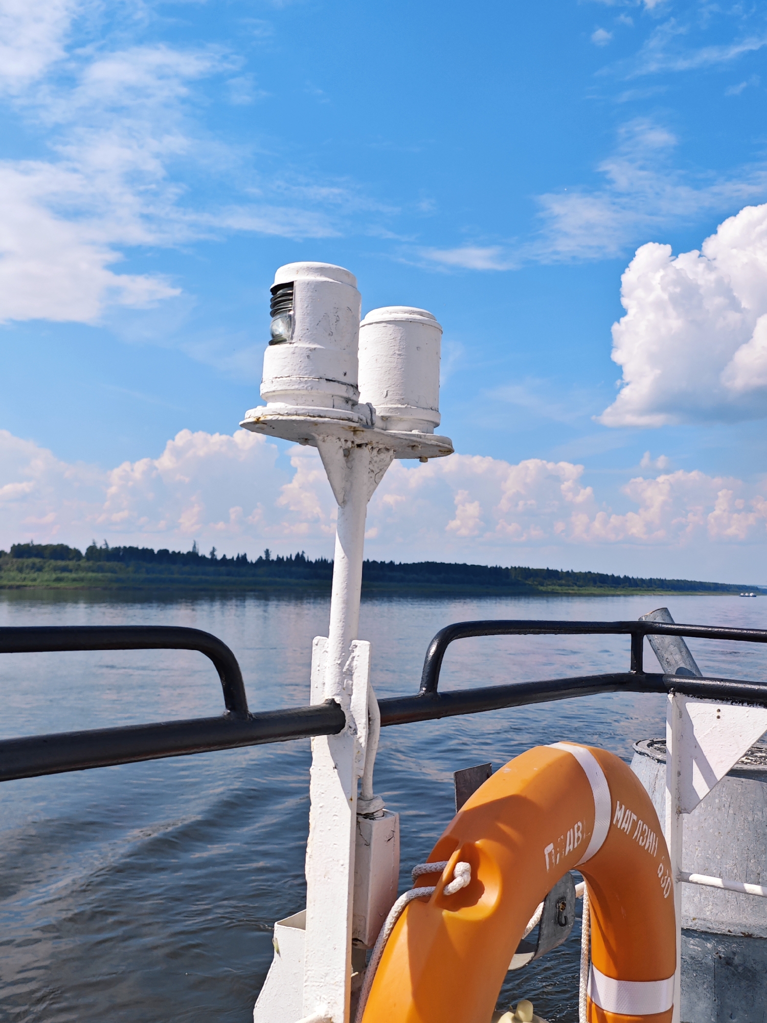 The image shows a view from the deck of a boat on a body of water, likely the Podkamennaya Tunguska River in Russia, on a sunny day. The sky is blue with scattered white clouds. In the foreground, there is a white railing and a white pole with two white cylindrical objects on top, possibly navigation lights. To the right, an orange life preserver is attached to a railing. In the background, a lush green forested shoreline is visible across the water. The water is calm with ripples. There are no people visible in the image.