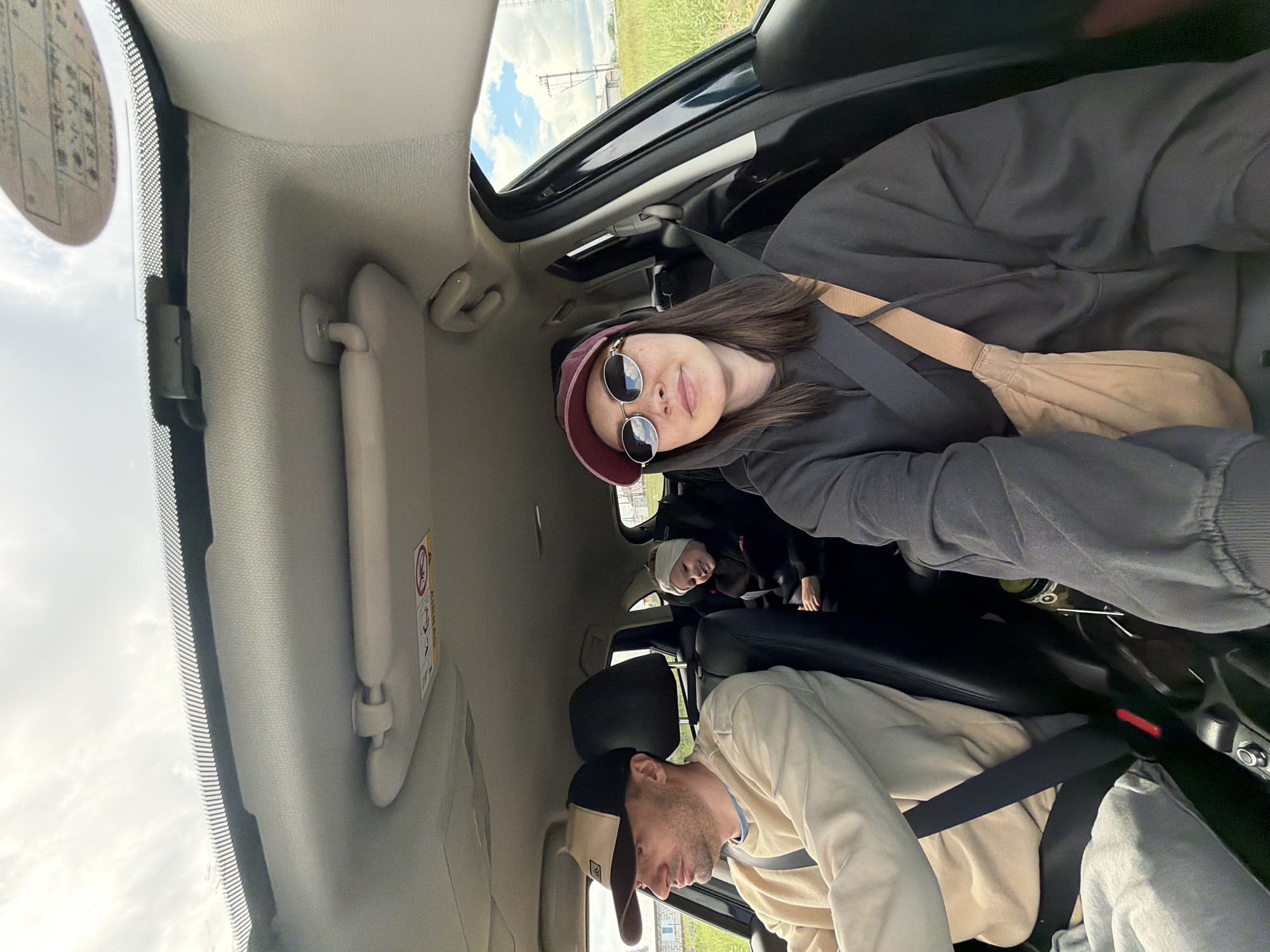 A vehicle interior, likely a car, is viewed from the front passenger seat at an unusual angle, suggesting the camera was tilted. In the front passenger seat, a man with a beard and wearing a light-colored shirt and a baseball cap is visible, secured with a seatbelt. In the back passenger seat, a woman with long dark hair, wearing sunglasses and a burgundy hat and a black hoodie, is also secured with a seatbelt and a tan bag. There are other people further back in the car. The vehicle has a sunroof open, revealing a view of a grassy field and cloudy sky. The location is the city of Chaikovsky, Russia. The car is in motion.