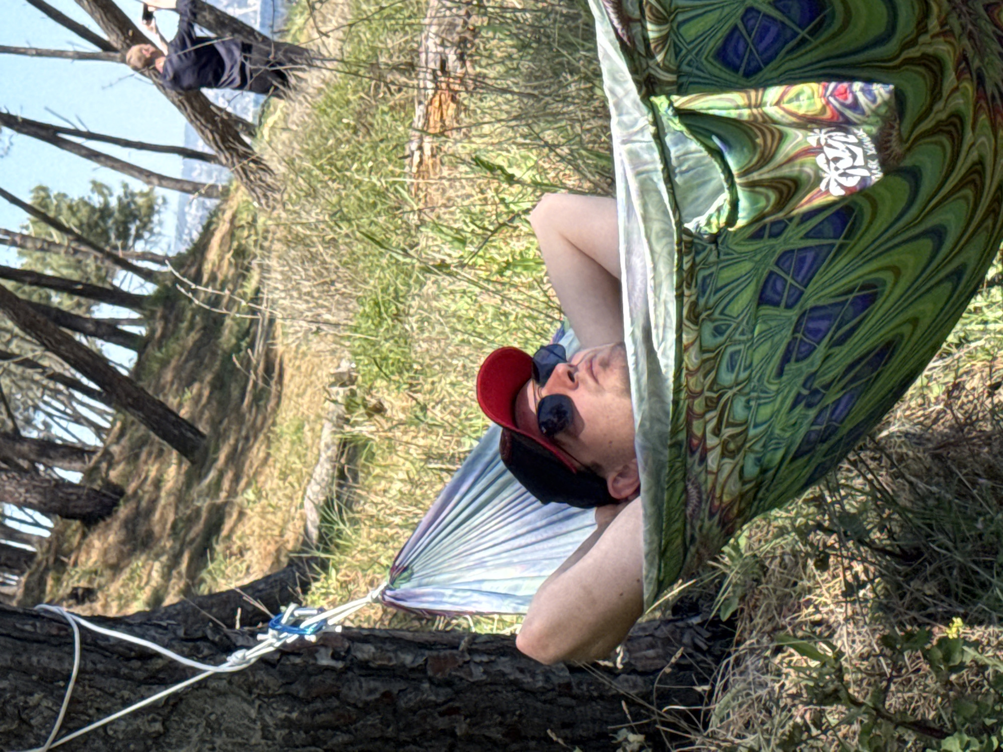 A man wearing a red baseball cap and sunglasses reclines in a patterned hammock strung between trees in a wooded area. Another person stands near trees in the background. The location is in a wooded area that appears to be on a hillside. The location is Varna, Bulgaria. The image appears to have been rotated.