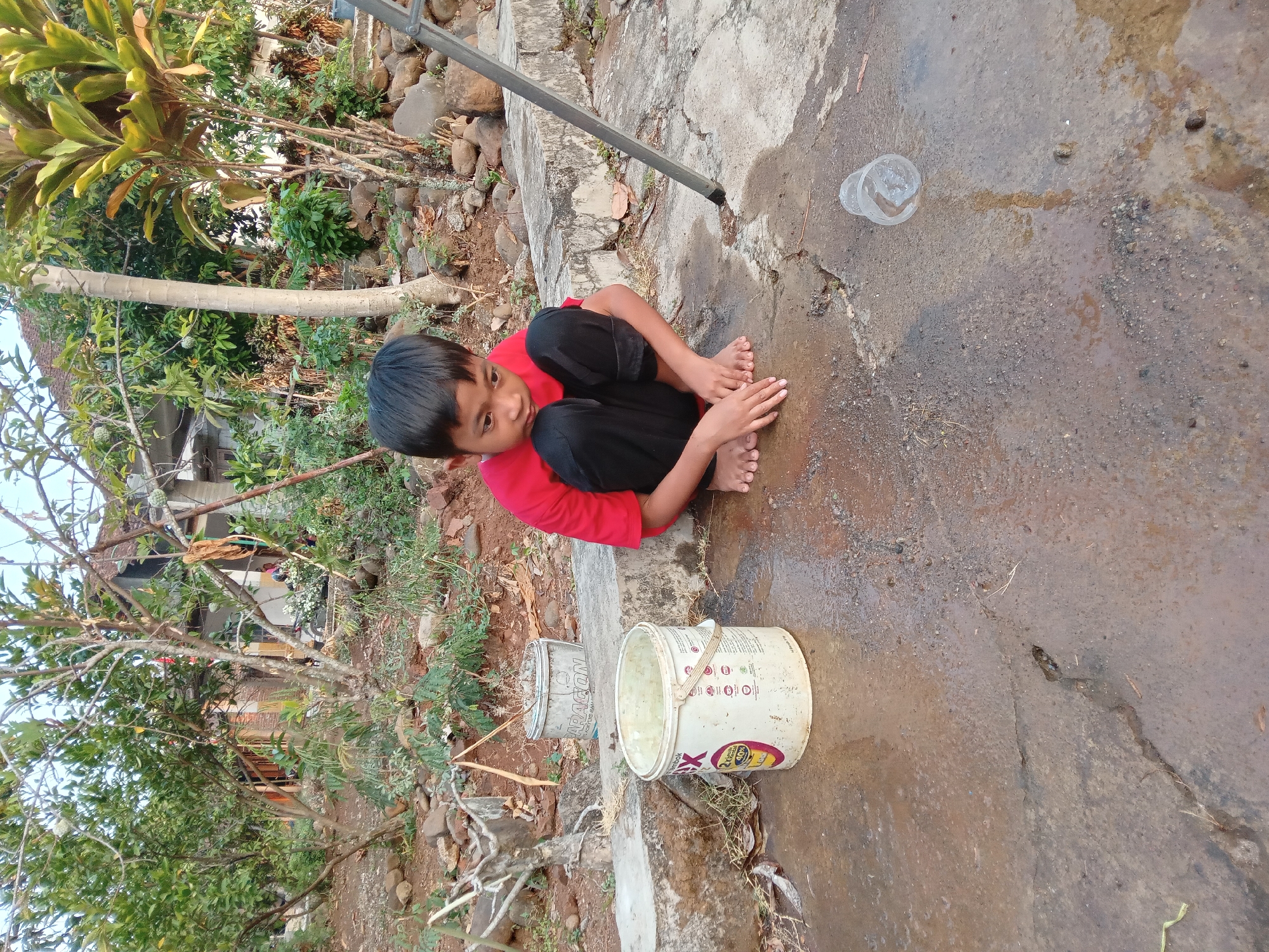 A young boy is crouching on a wet, cracked concrete surface in what appears to be a yard with dense vegetation and some buildings in the background. He is wearing a red t-shirt and black pants. In front of him is a white bucket with yellow and red lettering. To his right, there is a clear plastic cup lying on its side. A metal pole is visible on the right side of the frame. The ground is wet and appears to have some debris, including leaves and small stones. The overall lighting suggests it is daytime, possibly late afternoon due to the angle of the sun. The environment is outdoors, with trees, plants, and rocks visible in the background, suggesting a rural or semi-rural setting, potentially in Kedunggudel, Indonesia, as indicated by the location.