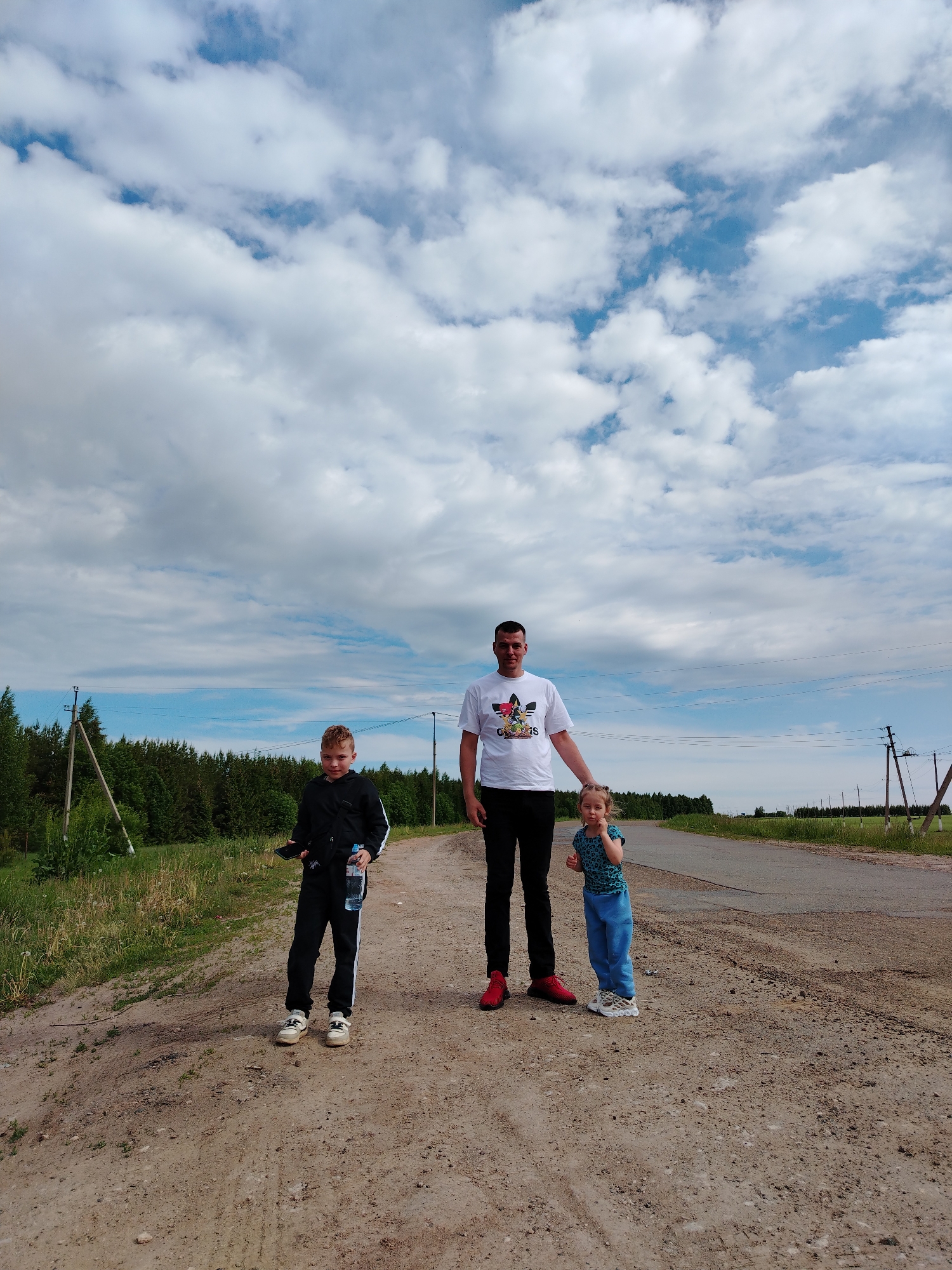 The image shows a man and two children standing on a dirt road. The man is wearing a white t-shirt, black pants, and red sneakers. He has his hand on the head of a young girl to his right. The girl is wearing a blue patterned shirt and blue pants, and white shoes. To the left of the man is a boy wearing a black tracksuit, and white shoes. He is holding a water bottle and a cell phone. The background is a cloudy sky and a green landscape with trees and telephone poles. The location appears to be Tavlarovo, Russia, based on the presence of Russian-style telephone poles and the rural landscape.