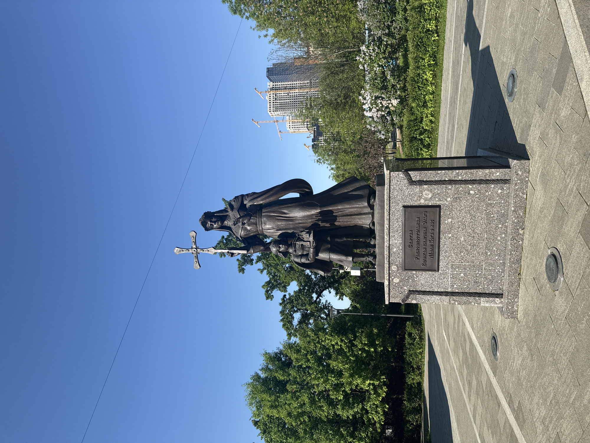 The image shows a bronze statue of Princess Olga, holding a cross, with her son Svyatoslav, at Borovitskaya Square in Moscow, Russia. The statue stands on a granite pedestal with a plaque in Russian. Behind the statue are green trees and bushes. In the background, modern buildings under construction can be seen. The sky is clear and blue. The statue is placed on a paved area with embedded lights.