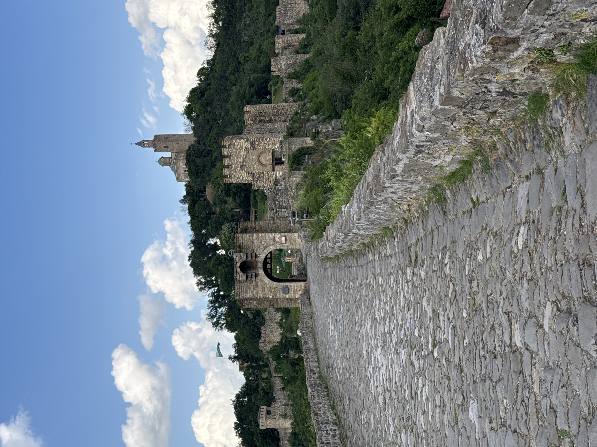 The image shows Tsarevets Fortress in Veliko Tarnovo, Bulgaria. The perspective is from a cobblestone path leading towards a large stone gate. In the background, a tall church tower rises above the fortress walls. The walls and towers are constructed of stone and are overgrown with vegetation. The sky is blue with scattered white clouds. A green flag flies near one of the fortress walls.