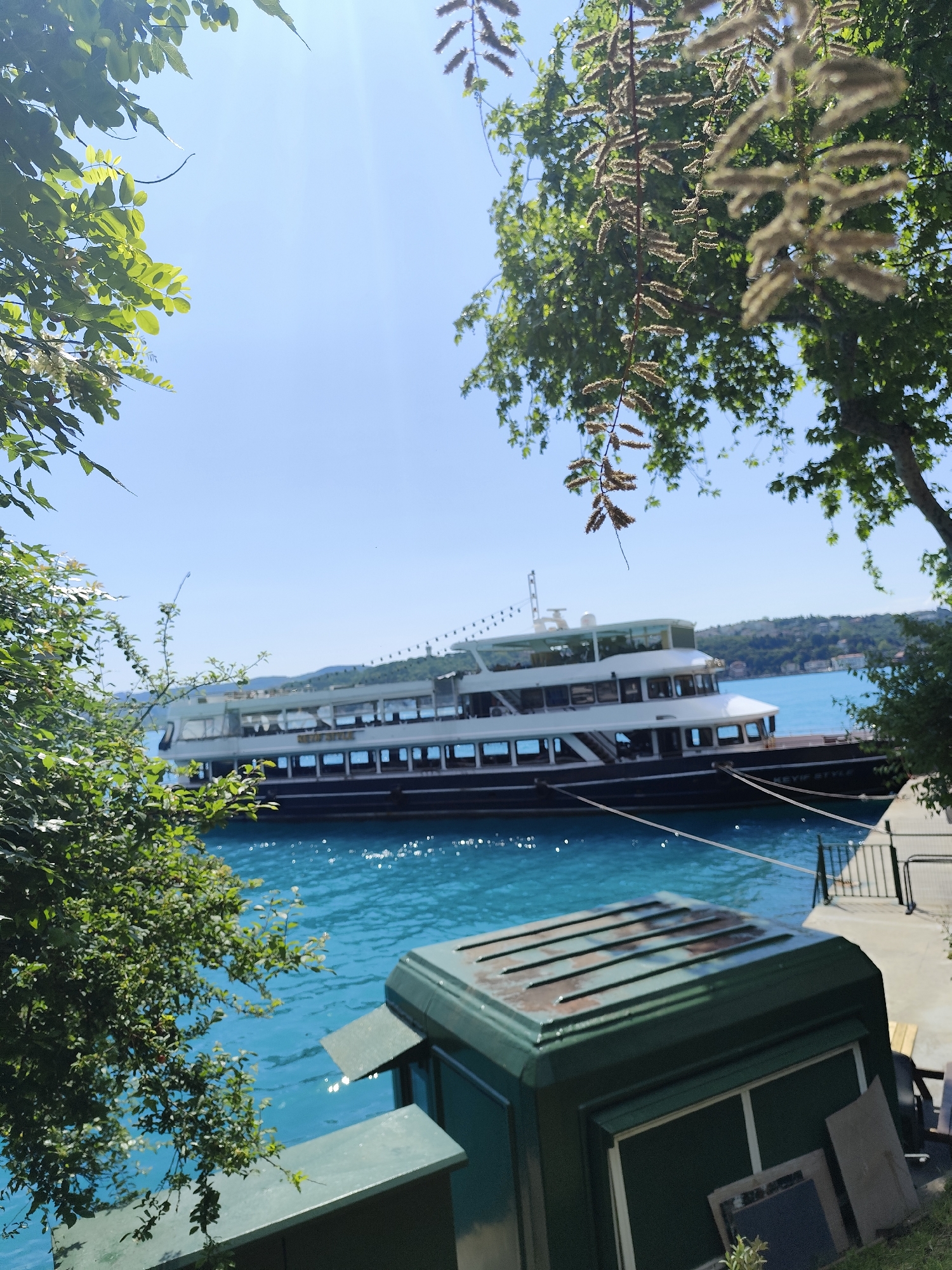 A partially-obscured, three-decked, passenger vessel rests on an aqua-blue body of water, likely the Bosphorus Strait, in Sarıyer, Turkey. The ship, named "Beyyip Style", is docked next to a concrete platform bordered by a metal railing. In the foreground, a dark green structure with a flat, ridged roof partially conceals the view. Trees and foliage frame the scene on both sides. The sky is a clear, light blue.