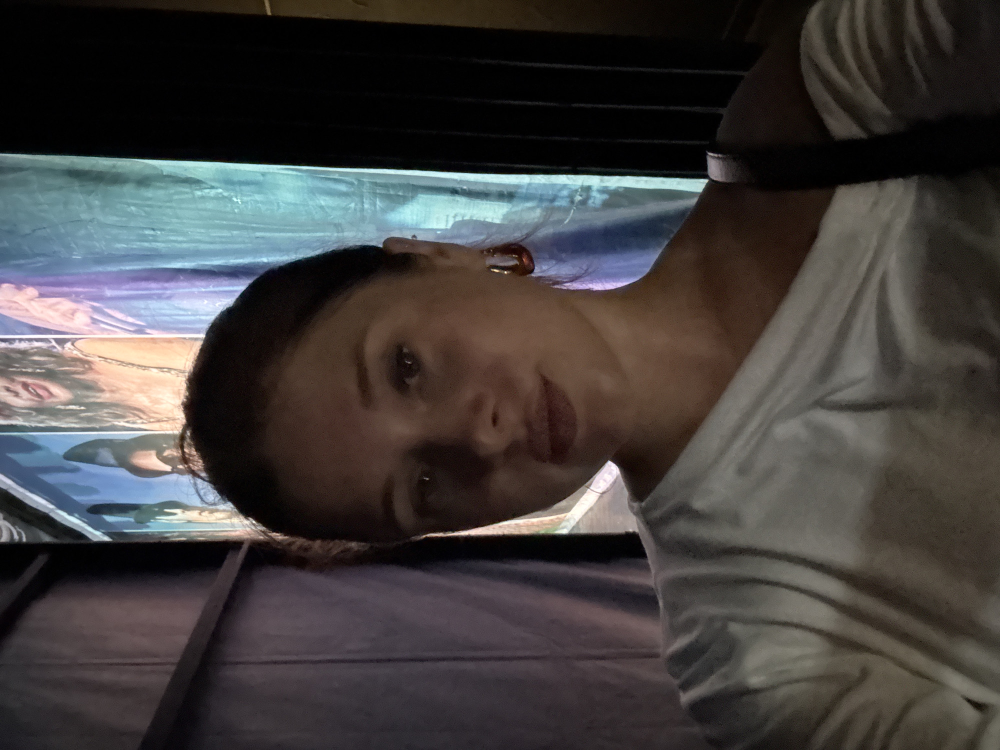 A young woman with brown hair is taking a selfie in a dimly lit hallway. She is wearing a white off-the-shoulder shirt and large hoop earrings. Behind her, a vibrant backdrop featuring images of people is visible. The lighting is low, casting shadows across her face and the surroundings, creating a dramatic effect. The setting appears to be indoors, possibly backstage or in a venue with decorative displays. There is no indication of the specific location being Aksu, Turkey.