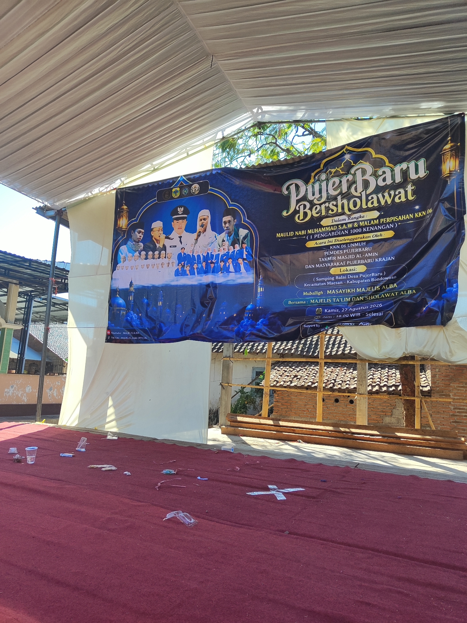 The image depicts an outdoor event space covered with a large, maroon-colored carpet. A banner with blue and gold lettering, titled "Pujer Baru Bersholawat," is prominently displayed. The banner features images of men in traditional attire and a mosque. Scattered across the carpet are various disposable plastic cups and containers, suggesting a recent gathering. In the background, a brick building and wooden structures are visible, indicative of a rural or semi-urban setting. The event appears to be held under a white tent. The text on the banner indicates the event is in Pujer Baru, Indonesia, and is scheduled for Kamis, 27 Agustus 2026.