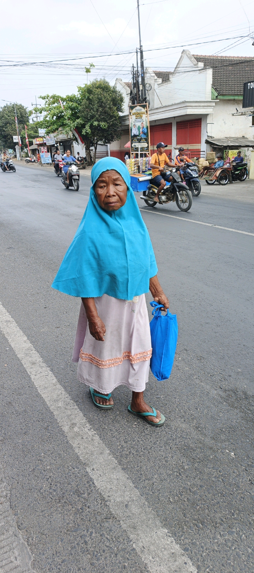 An elderly Indonesian woman wearing a bright turquoise hijab and a light pink skirt walks along a paved road in Kasiyan, Indonesia. She is holding a bright blue reusable bag in her right hand and wears teal flip-flops. The road is asphalt, marked with a white line. In the background, several motorcycles are visible, with riders on them. Buildings with red accents line the street, and there are trees and utility poles. The sky is overcast.
