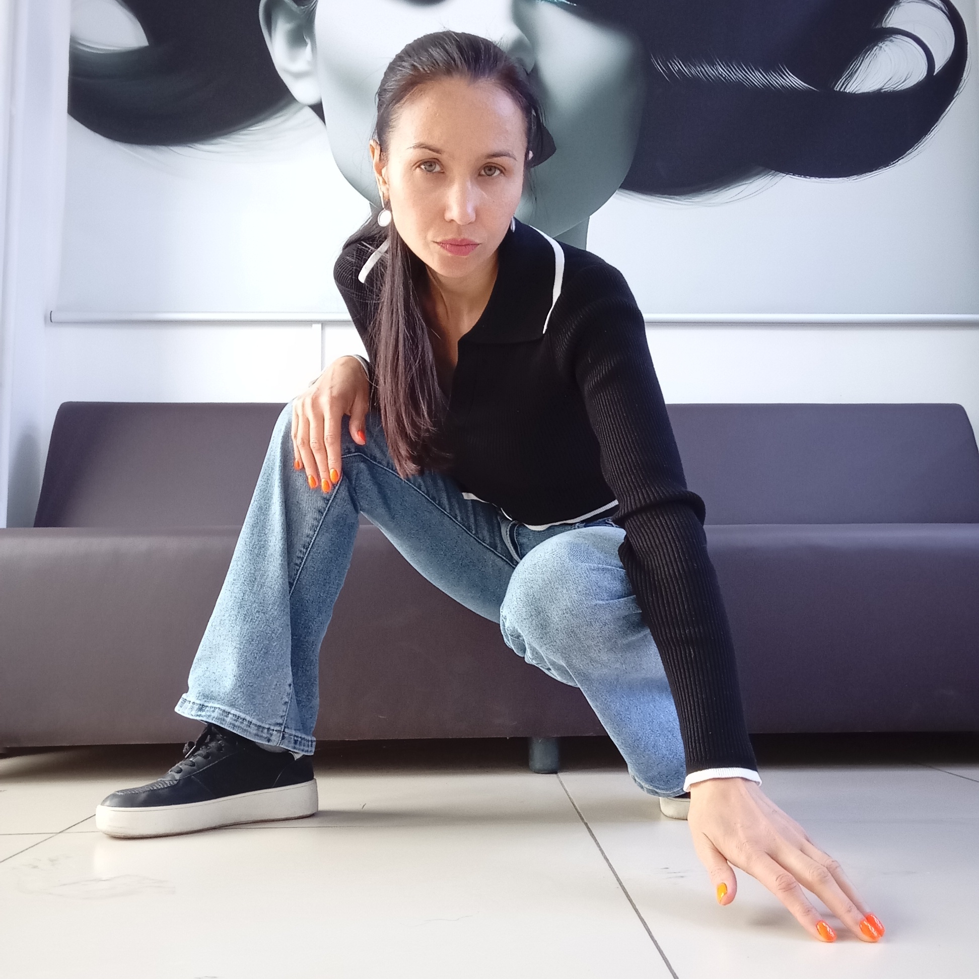 A woman is crouching indoors in Izhevsk, Russia. She is wearing blue jeans, black sneakers with white soles, and a black ribbed long-sleeved top with white accents at the collar and cuffs. She has long dark hair pulled to one side, and orange nail polish. Behind her is a grey couch and a large black and white print of a woman's head. The floor is white tile. She is looking directly at the camera with a neutral expression, with one hand resting on her knee and the other extended towards the floor.