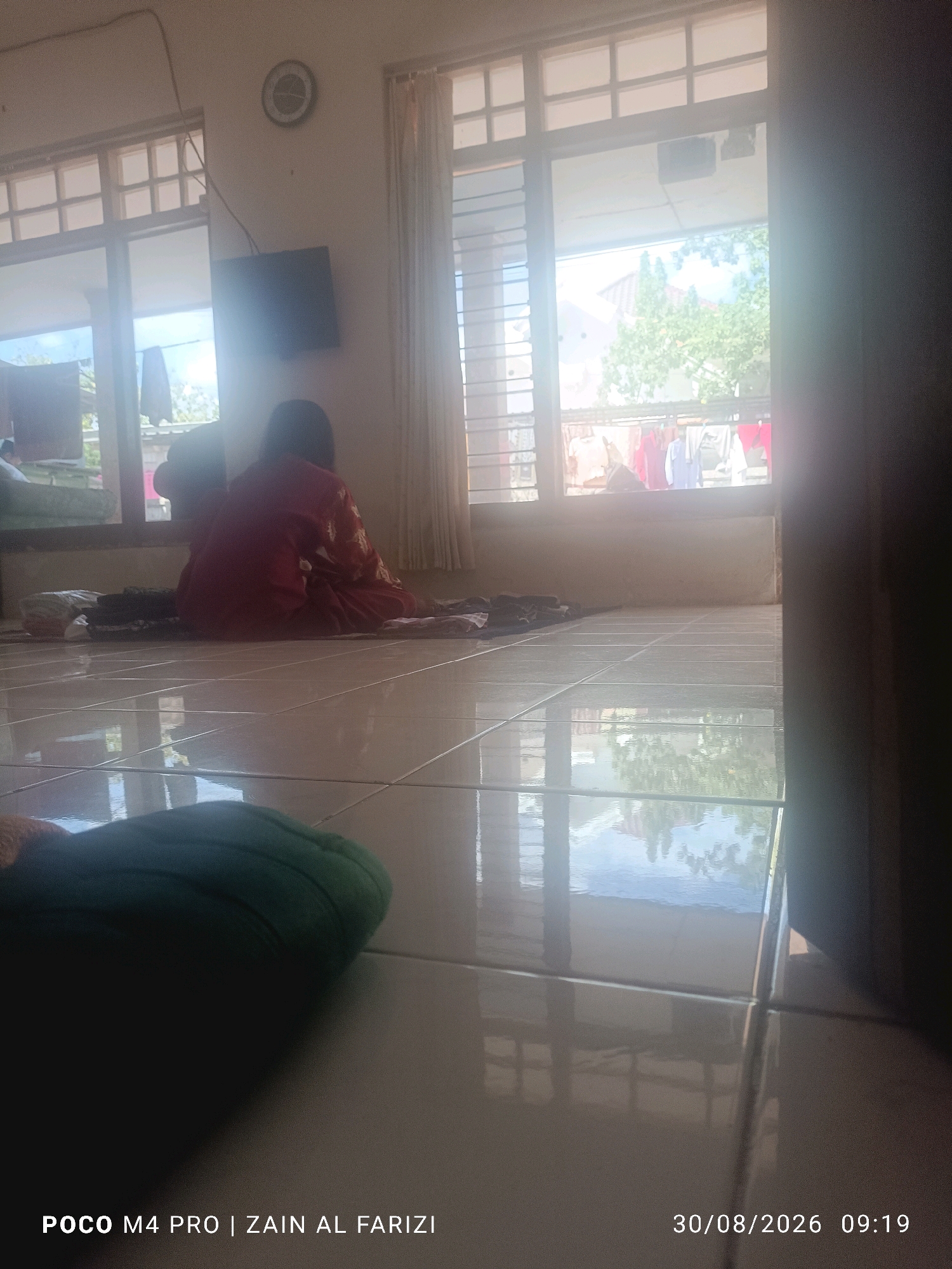 A person wearing a red garment is seated on the floor in front of a window. The floor is tiled and reflective. A clock is visible on the wall above the window. Laundry is hanging outside the window. The image also contains text overlays indicating the device used, the photographer's name, and the date and time. The scene is likely indoors, possibly a residential setting, in Karang Pao, Indonesia, given the metadata.