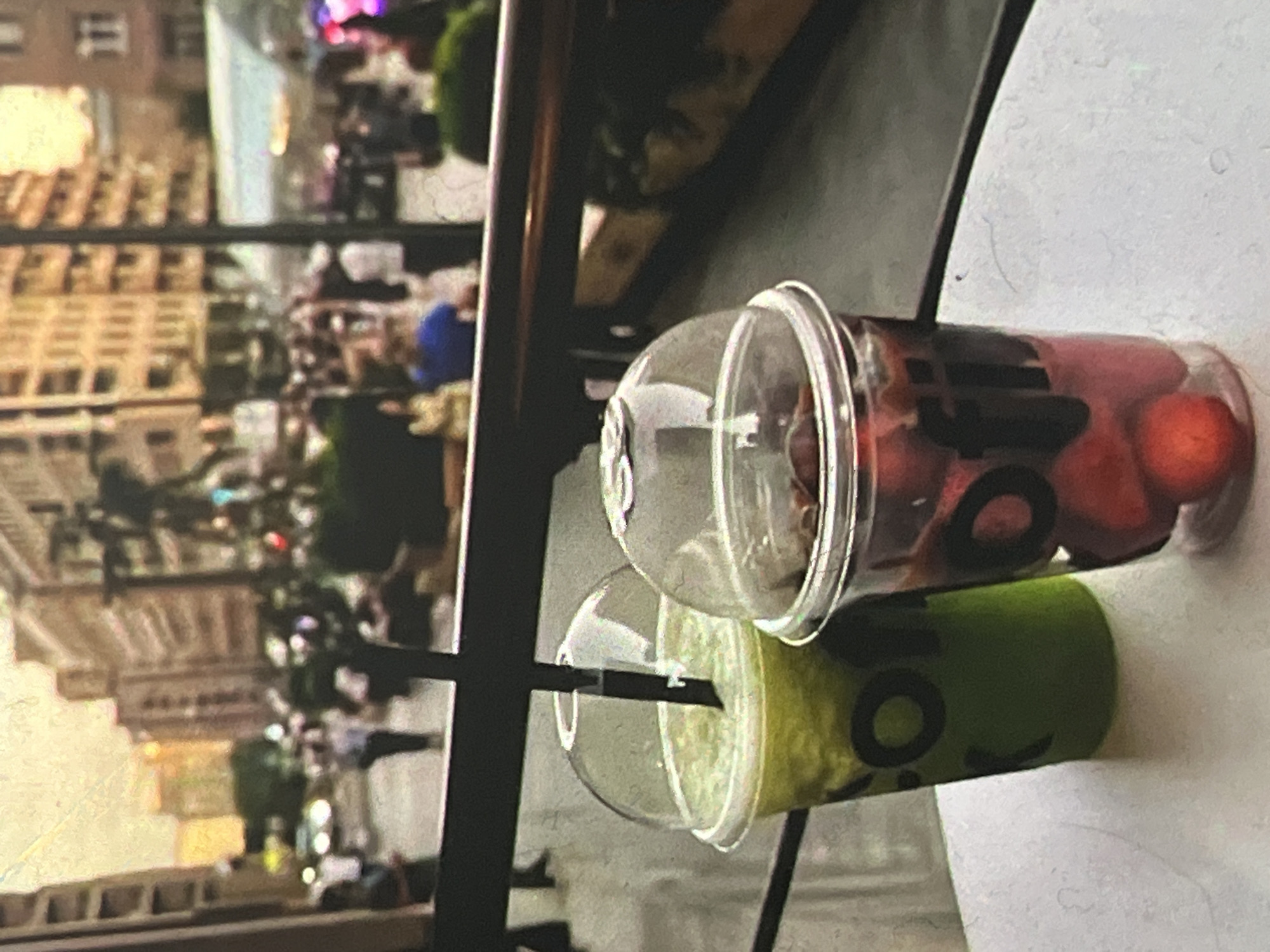 This is a medium shot, taken from a slightly elevated angle, of two clear plastic cups with domed lids, filled with beverages and straws, sitting on a white surface. The cup on the left contains a green, blended drink. The cup on the right contains a dark red liquid with visible whole red fruits, possibly strawberries or cherries, submerged within it. Both cups have dark lettering on them, which appears to be a logo.
In the background, the scene is blurred but suggests an outdoor urban environment. Buildings with multiple stories and windows line a street, with people and vehicles visible in the distance. The lighting indicates it is daytime, possibly late afternoon or early evening, with a warm glow to the sky. The overall impression is that of a cafe or dining area overlooking a busy city street.
The image does not provide any direct visual cues to confirm its location as Krasny Yar, Russia.
