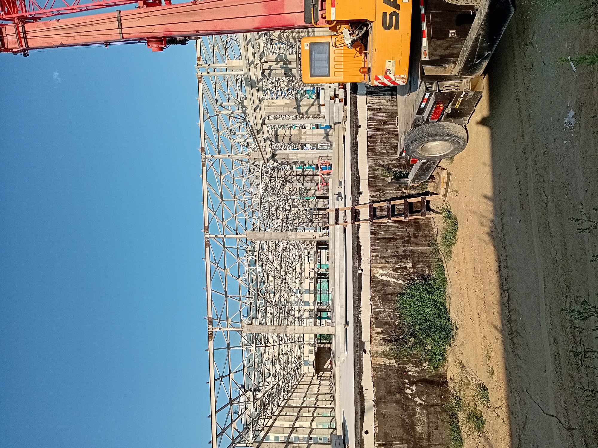 An outdoor, daytime, wide-angle shot captures a construction site in Semey, Kazakhstan. The primary focus is on the skeletal framework of a large building under construction, composed of numerous steel beams and concrete pillars. The structure appears to be a warehouse or industrial facility, evidenced by its expansive open interior and the repetitive nature of its supports. In the foreground, a large, yellow crane is partially visible on the right side of the frame, its red boom extending upwards. Adjacent to the crane, a black trailer with a spare tire is parked. A wooden ladder leans against a concrete wall. The ground is a mixture of dirt and sand, with tire tracks imprinted in places. Green vegetation, including bushes and grass, grows sporadically around the base of the concrete wall and in the foreground. The sky is a clear, bright blue. The overall scene suggests an active construction project in progress.