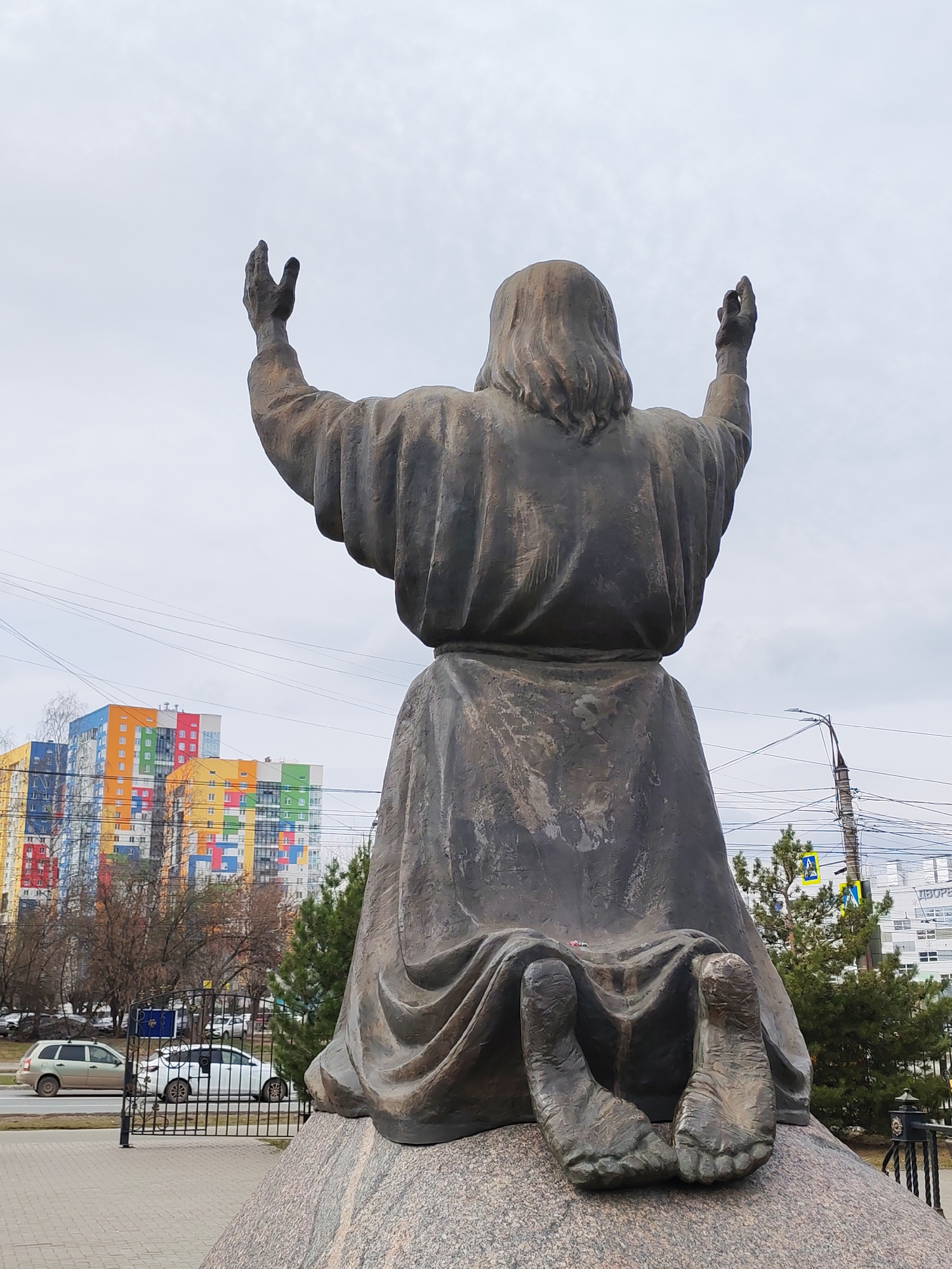 The image features a bronze statue, viewed from the back, depicting a figure kneeling on a rough stone base with arms raised towards an overcast sky. The figure, likely Jesus Christ, has long hair and wears draped robes, suggesting a pose of prayer or supplication.
In the background, an urban environment unfolds under a cloudy sky, indicating daytime. Dominating the midground are several modern, multi-story residential buildings distinguished by their vibrant, block-like color patterns in shades of red, orange, yellow, green, and blue. Bare trees and some evergreen bushes are scattered throughout the scene.
A road is visible in the middle ground, with cars, including a silver station wagon and a white sedan, parked or driving. A black metal fence separates the statue's immediate area from the road. Overhead, numerous power lines crisscross the sky. On the right, a street light pole and a yellow pedestrian crossing sign are visible. Faintly visible text on a white building in the far background appears to include the Russian word "ДВОР" (DVOR). The overall setting suggests a city street in Izhevsk, Russia, characterized by a mix of solemn statuary and modern, colorful architecture.