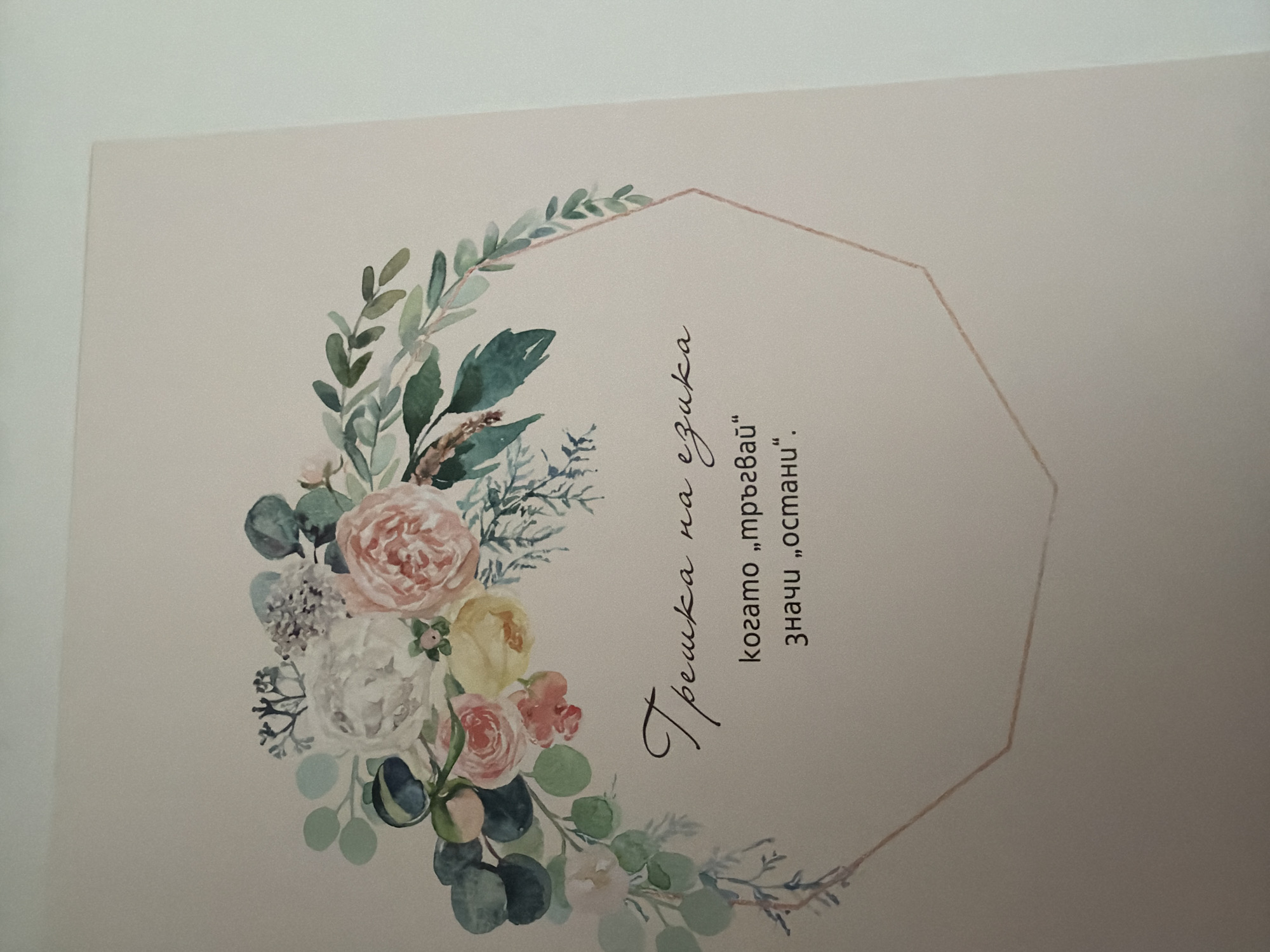 A floral watercolor design is printed on a light-colored surface. The design features a wreath of various flowers, including peonies and roses, in shades of white, pink, and yellow, interspersed with green eucalyptus leaves and other foliage. A thin, rose-gold colored hexagonal outline encloses a portion of the text. Below the floral design, in elegant script, are the words "Трешка на езика" (Treshka na ezika). This is followed by the phrase "когато „тръгвай“" (kogato "trugvai") meaning "when 'leave'". Below this, it reads "значи „остани“." (znachi "ostani".), meaning "it means 'stay'." This appears to be a decorative card or print. The image was captured in the city of Pazardzhik, Bulgaria.