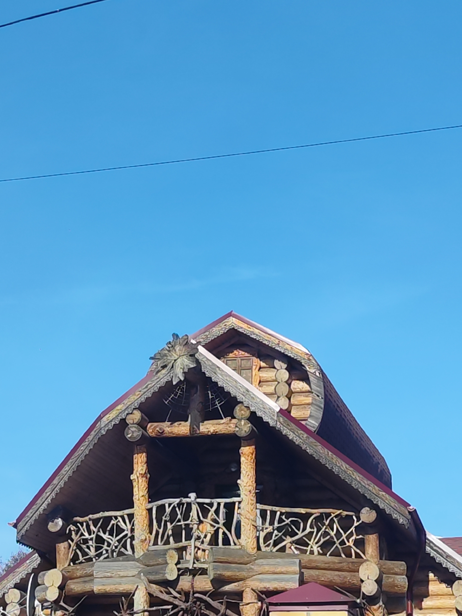 The image shows the upper portion of a two-story log building under a clear blue sky. The structure appears to be located in Chaikovsky, Russia based on information available in the description. The building features decorative carvings on its roofline and around the balcony. The balcony has a unique railing made from intertwined branches. A small window is visible near the roof's peak, and a decorative element is present at the top of the gable. An overhead wire is visible against the sky.
