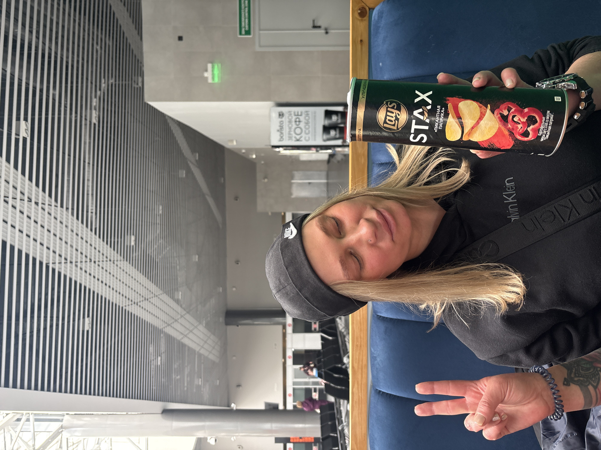 A sideways shot of a woman holding a tube of Lay's Stax "Spicy Paprika" potato chips while sitting in an airport or train station waiting area. The location may be in the city of Sokol, Russia, given the presence of Russian text in the background. The woman has fair skin, blonde hair, and wears a black beanie, a black hoodie with a Calvin Klein strap, and a dark bracelet. She holds the potato chip tube in her right hand and gives a peace sign with her left hand, which has a visible tattoo. The tube of chips has the Lay's logo, "STAX" text, and a picture of paprika-flavored chips. In the background is a sign advertising "Barista Zernovoy Coffee." The waiting area has metal grills for the ceiling, with a variety of seating areas and other people visible in the distance.