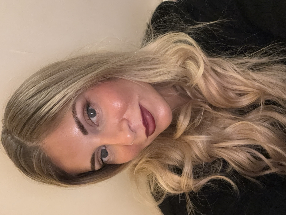 A fair-skinned woman with blonde, wavy hair looks at the camera. Her hair is parted in the middle. She wears dark lipstick. Her eyebrows are thick and well-defined. A black garment is visible behind the cascade of curls. The background appears to be a light-colored wall. The picture is oriented at an angle.