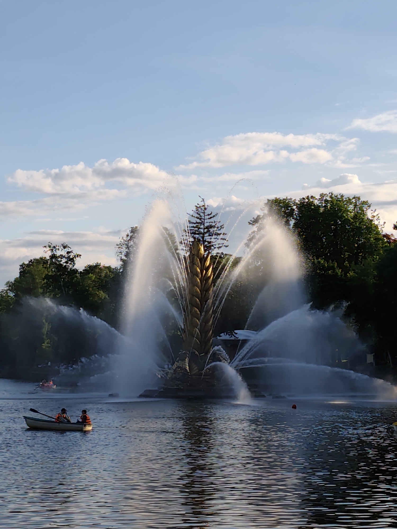 The image depicts a large fountain in a lake in Moscow, Russia. The fountain's central structure resembles a golden wheat sheaf, with water jets spraying outwards and upwards. Two people in a rowboat are visible in the foreground, wearing orange life vests and holding paddles. In the background, beyond the fountain, are trees and other people in rowboats. The sky is light blue with scattered white clouds. The water in the lake reflects the sky and the surroundings. A red buoy is visible in the water near the fountain.