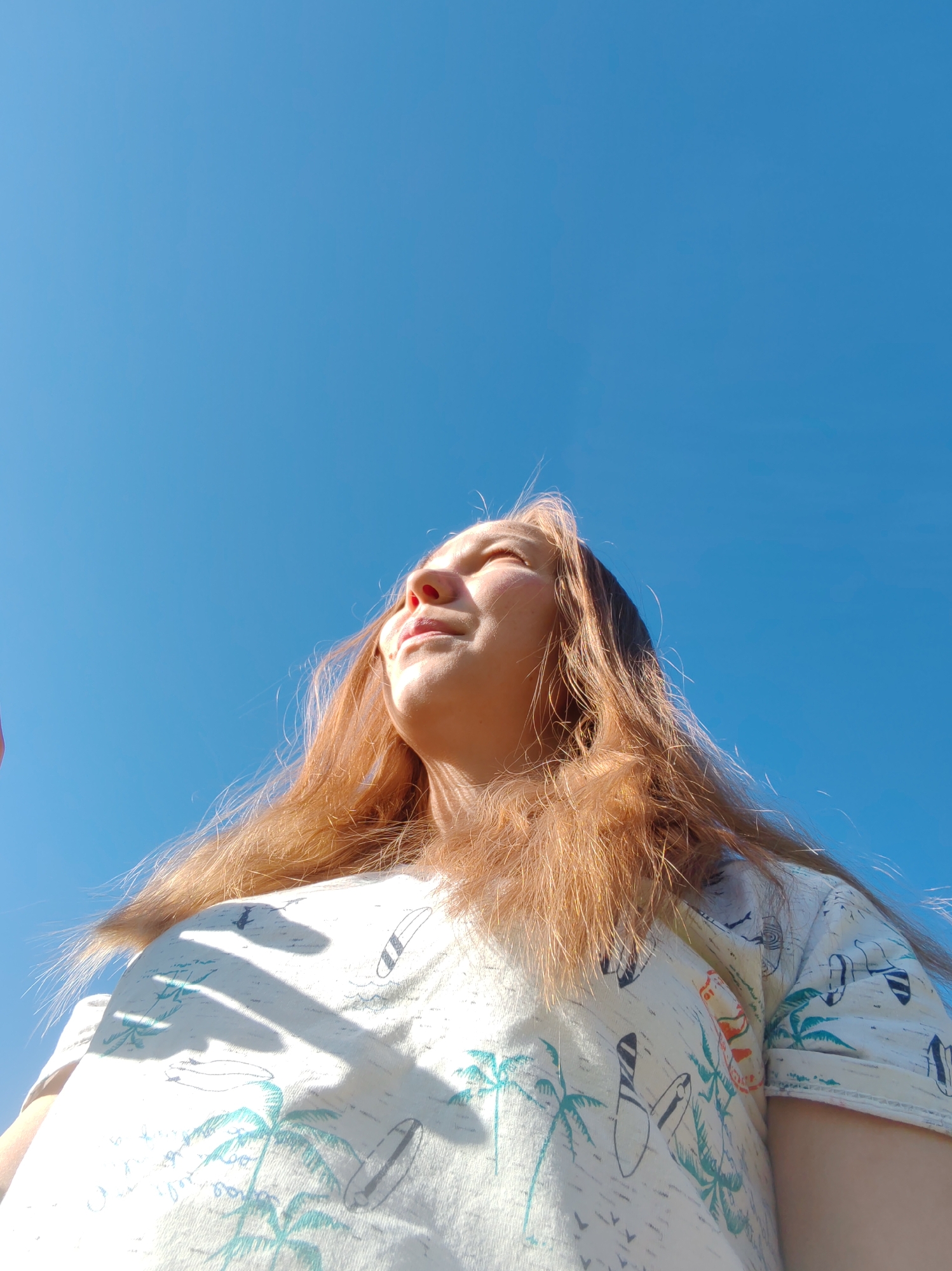 The image is a low-angle, close-up shot of a young woman with reddish-brown hair, looking upwards towards a clear blue sky. She is wearing a light-colored t-shirt with a repeated pattern of palm trees, surfboards, and small birds. Her face is illuminated by sunlight, with her eyes closed and a slight smile. Shadows from what appear to be fingers are cast across the front of her t-shirt. The background is entirely the bright blue sky, suggesting an outdoor setting during daylight. There are no visible landmarks or contextual elements to place this image specifically within the city of Чухлэм, Russia.