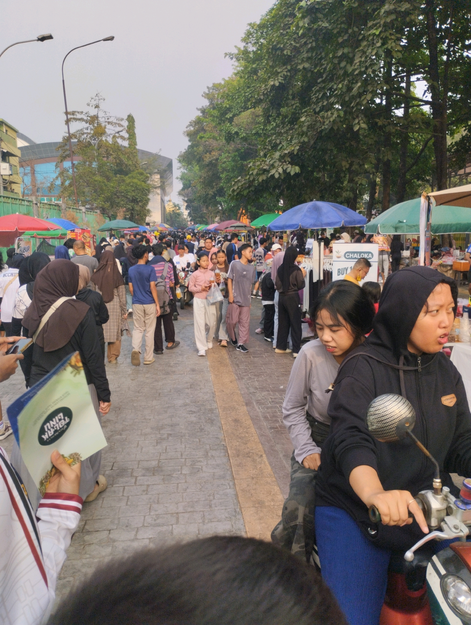 This is a street-level, eye-level, full shot of a busy outdoor market in the city of Bekasi, Indonesia. The scene is bustling with activity, suggesting a daytime event. A large crowd of people is walking along a paved walkway, which is lined with various market stalls and vendors. The stalls are covered with umbrellas of different colors, including red, green, and blue. In the background, there are buildings and trees, indicating an urban environment.
Several individuals are captured in various poses and interactions. In the foreground, a person on a motorcycle is visible, with a woman in a black hoodie and blue pants riding on the back. The woman on the back appears to be looking towards the right, with her mouth slightly open. Another woman, wearing a grey long-sleeved shirt, is seated in front of her on the motorcycle.
Further into the market, people are browsing goods, talking, and walking. Some individuals are wearing headscarves, which is common in Indonesia. One person is holding a leaflet with text in what appears to be Indonesian. The overall impression is that of a vibrant community gathering or shopping event. The lighting suggests it is either early morning or late afternoon due to the soft, diffused light. The sky is overcast, contributing to the muted tones of the scene. The specific location is implied to be Bekasi, Indonesia, as per the prompt, though no specific landmarks are identifiable from the image itself that definitively pinpoint a particular spot within the city.